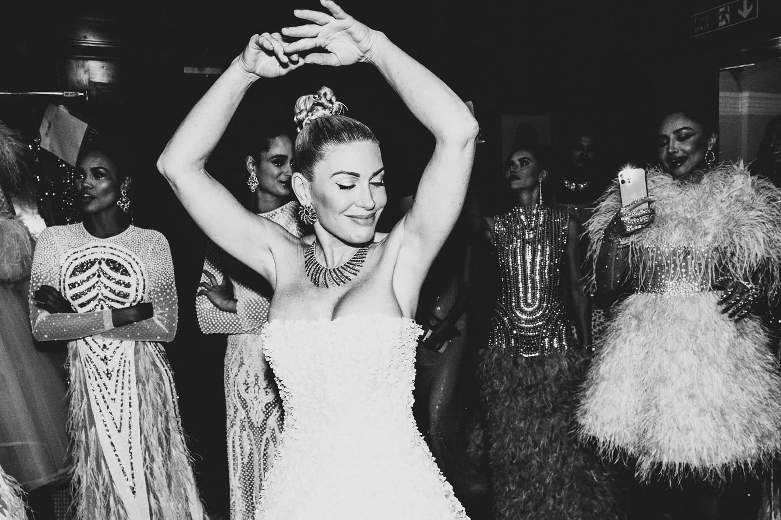 A black-and-white backstage scene at a fashion show featuring several models wearing ornate, embellished dresses with feathers and intricate beading. One central figure has her arms raised above her head in a graceful pose, eyes closed with a soft smile, wearing a strapless textured gown and a statement necklace. Other figures stand behind, some blurred and some illuminated by a phone light in the dimly lit environment.