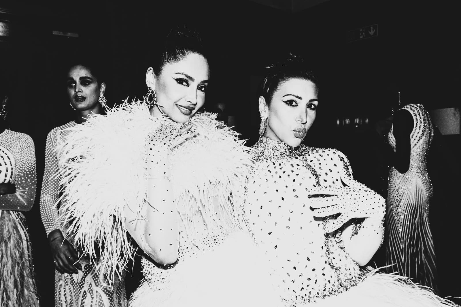 A black-and-white backstage medium shot showing two individuals posing closely. The person on the left wears a white feathery garment with delicate embellishments and large ornate earrings. The person on the right is pouting toward the camera, wearing a high-necked, heavily sequined dress and matching gloves. Other figures in embellished garments are visible in the dimly lit background.