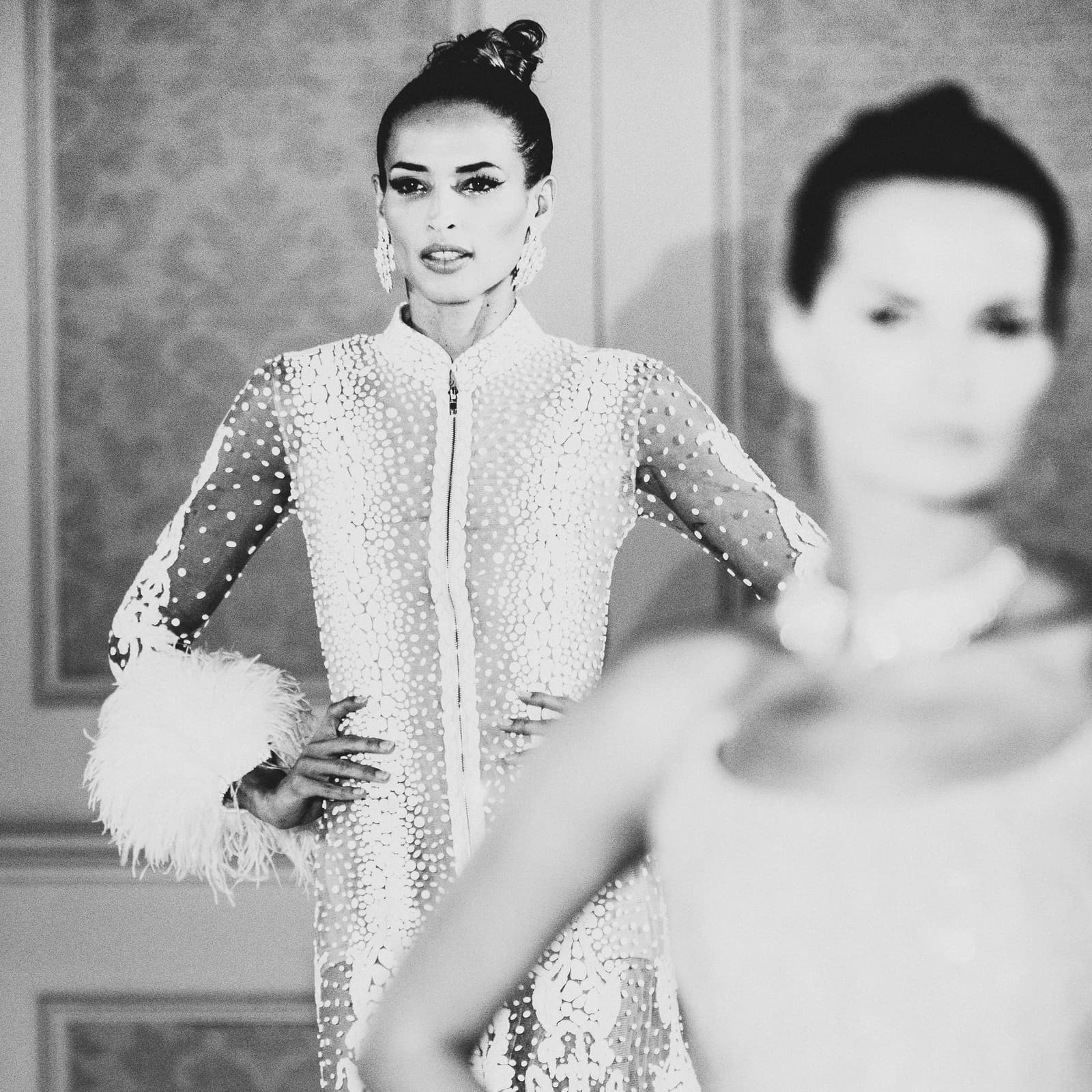 A black-and-white medium shot of a model on a runway, wearing a high-necked, sheer, heavily embellished gown with feather-trimmed cuffs. Her hands are on her hips, and she wears large statement earrings. In the foreground, another model is visible but out of focus, blurred by shallow depth of field.