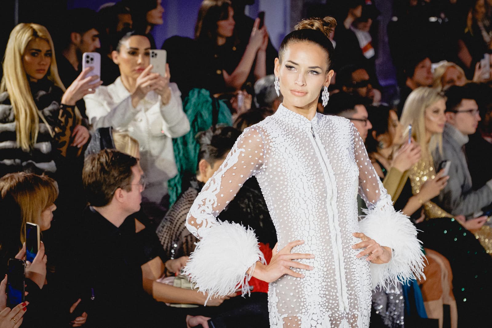 A model walks a catwalk in a white sheer dress adorned with intricate beadwork and long white feather cuffs at the wrists, her hair pulled back in a high bun. She is surrounded by a seated audience who are holding up smartphones to capture the moment, with bright studio lighting reflecting on the scene.