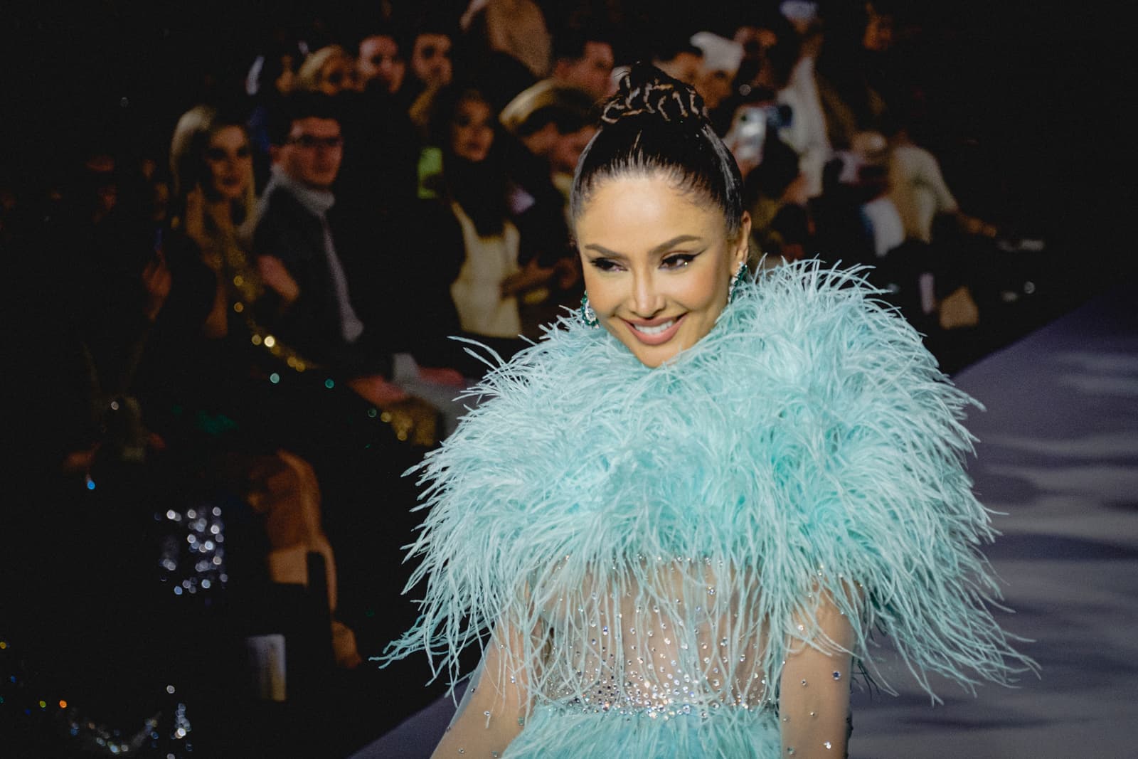 A model walks a catwalk wearing a light blue garment covered in abundant feathers around the shoulders and torso, over a sheer bodice embellished with silver crystals. The hair is pulled back into a high, sleek bun. The background shows a blurred audience seated in low light.