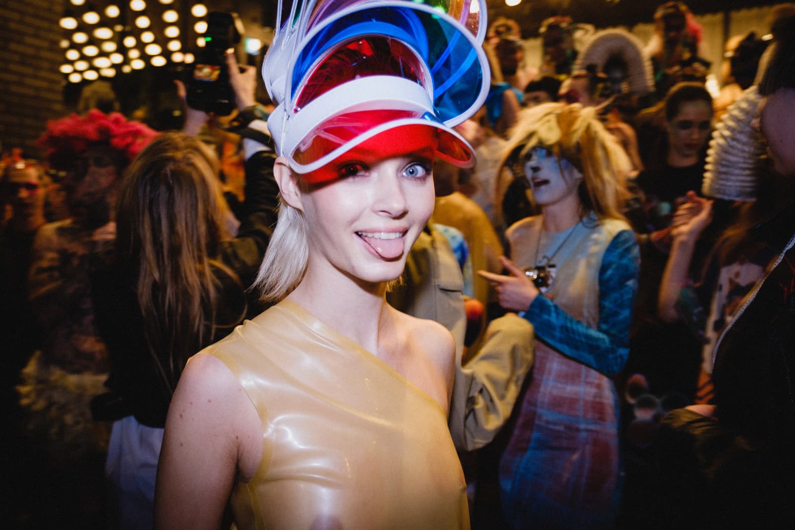 A close-up shot of a person facing the camera, wearing a translucent, pale, one-shoulder latex top and a multi-coloured, layered plastic visor. The person is sticking their tongue out with a playful expression. The background is a crowded backstage area at a fashion show, filled with other people in stylized outfits, warm lights, and soft-focus silhouettes.