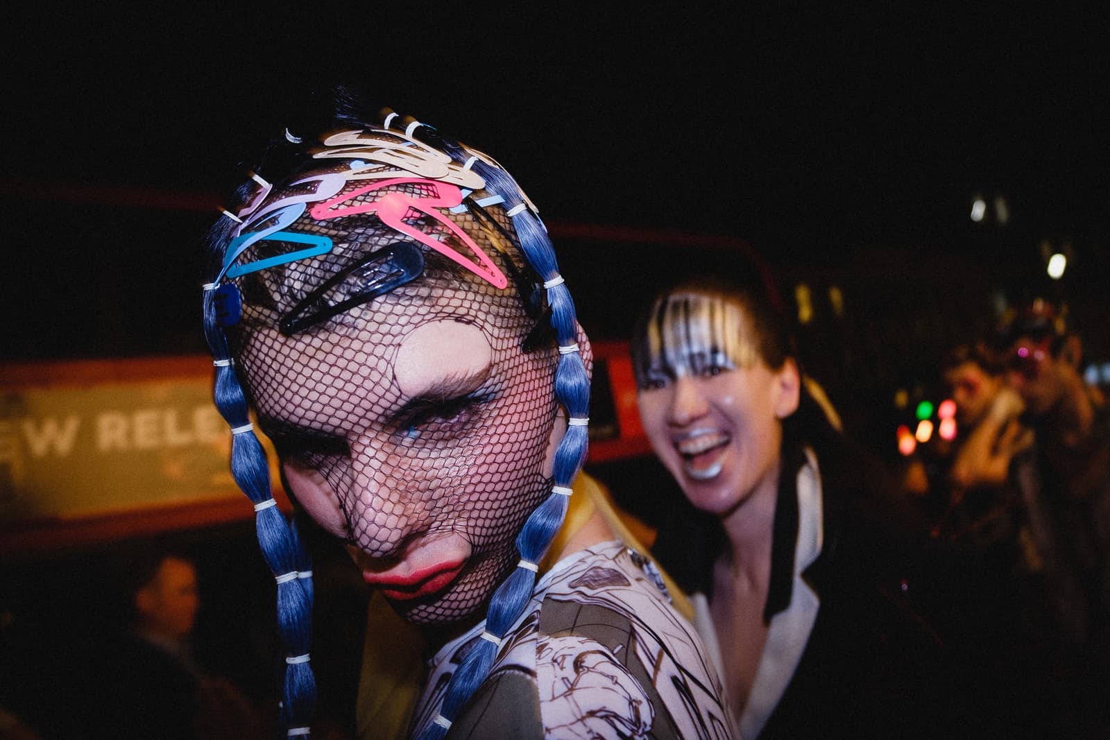 Close-up portrait of a person wearing a black fishnet mask over their face, adorned with bright multi-coloured hair clips and blue braided hair, standing in front of another person who is laughing with a bright silver lip application. The setting appears to be a dimly lit backstage area.
