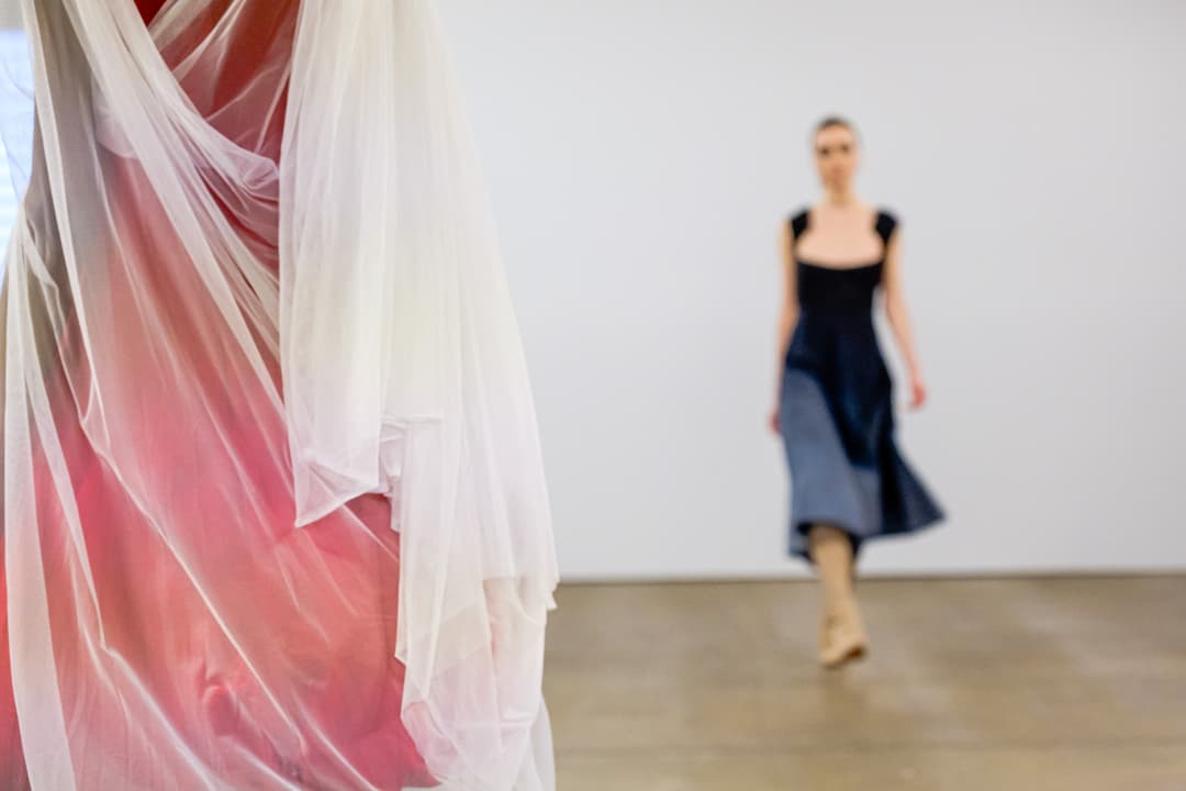 A close-up of translucent, layered white fabric draped over a red garment dominates the left foreground. In the soft-focus background, a person walks down a white-walled catwalk wearing a sleeveless dark dress with a square neckline and a full skirt, paired with light-coloured knee-high boots.