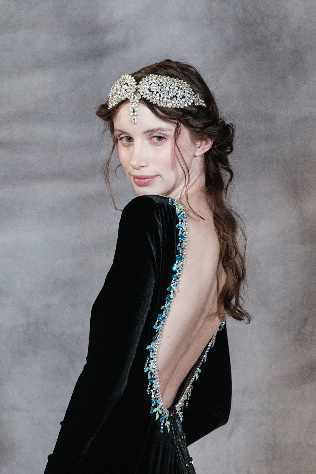 A person with long brown hair styled in a loose updo, wearing an ornate, silver-toned crystal tiara across the forehead, is captured in a medium shot from behind, looking over their shoulder toward the camera. They are dressed in a long-sleeved, black velvet gown featuring a deep, open back embellished with intricate blue, teal, and silver beadwork along the spine. The background is a plain, mottled grey wall illuminated by soft, even studio lighting.