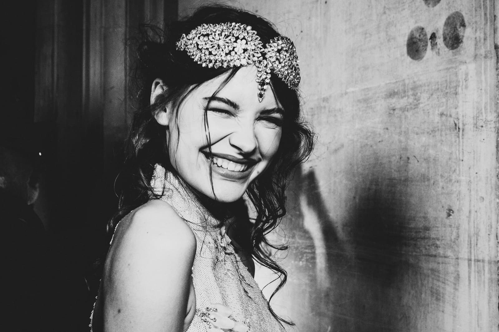 A black-and-white medium shot of a laughing person wearing a large, ornate, sequined headpiece across their forehead. The person has dark hair and is wearing a textured sleeveless top, captured in sharp focus against a plain, light-colored wall with visible texture.