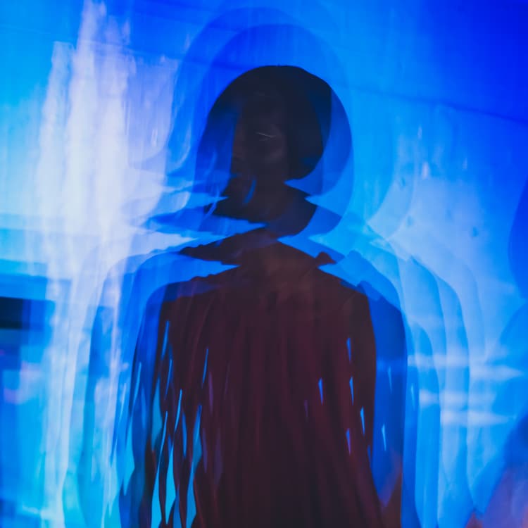 A long-exposure photograph of a figure in motion, bathed in deep blue light. The subject is captured in multiple overlapping silhouettes, creating a ghostly, blurred trail of movement against a plain blue background.