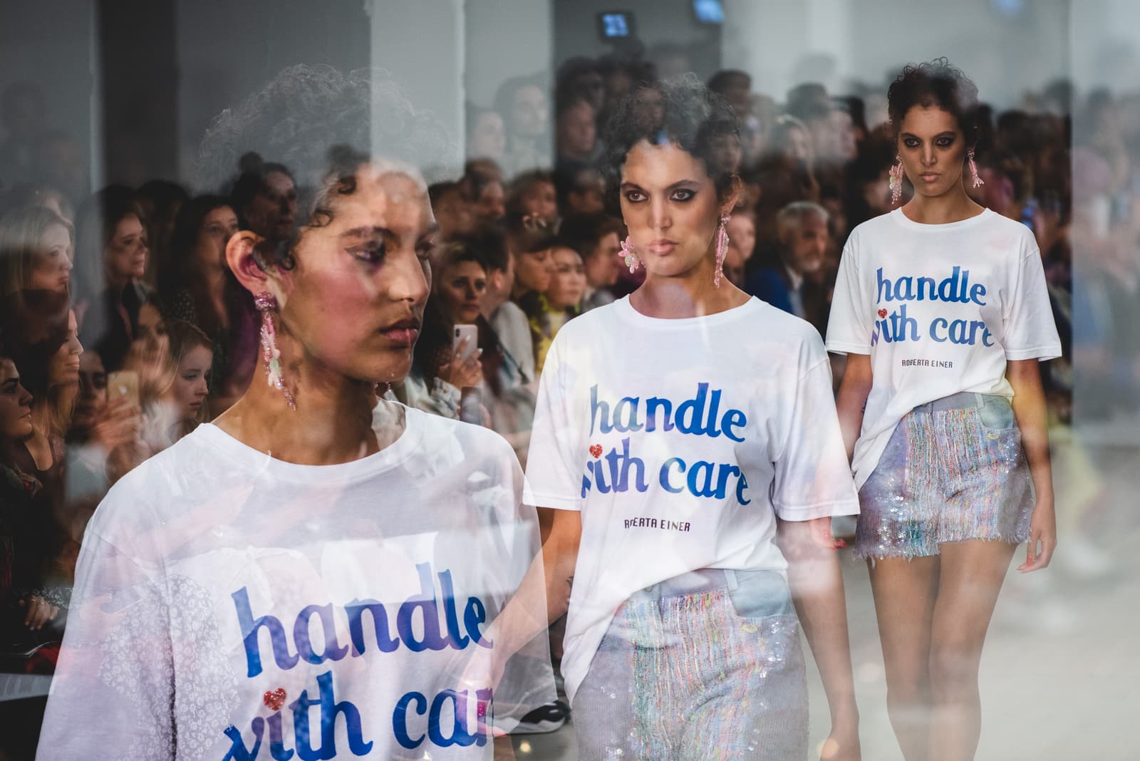 A multiple exposure shot shows three overlapping views of a model walking down a runway. The subject is wearing a white t-shirt with the text 'handle with care' printed in blue and sparkly shorts, accented with long, shimmering fringe. The background is filled with blurred audience members holding smartphones and cameras.