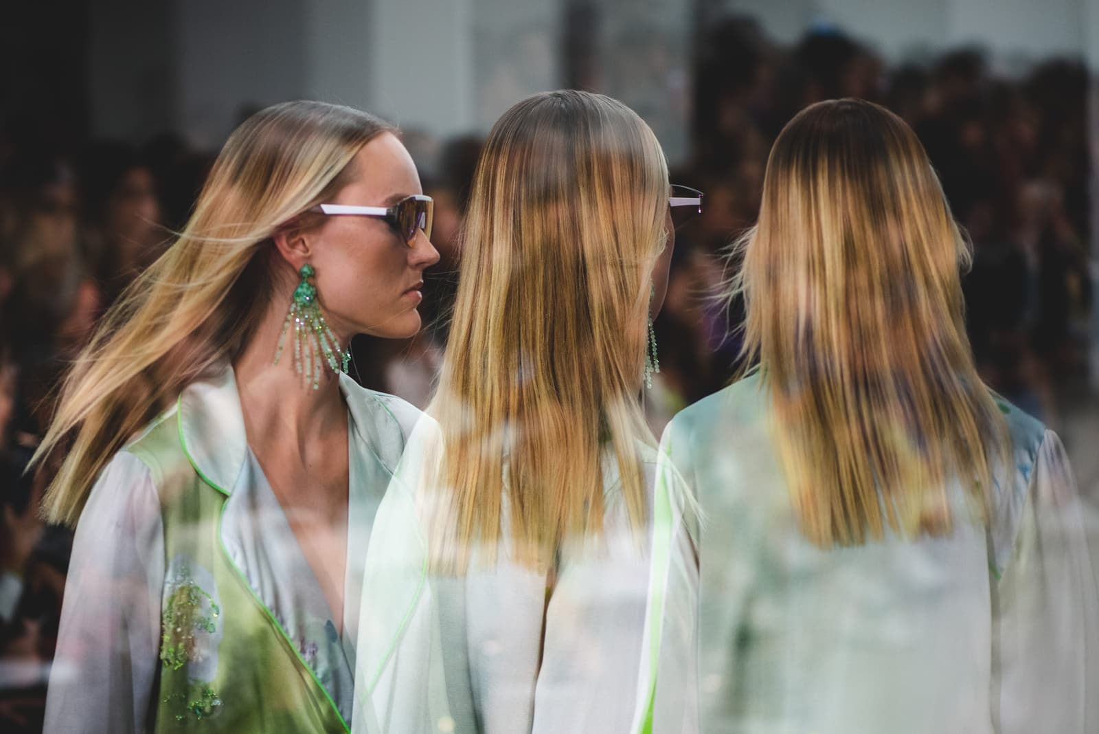 A multiple exposure image shows three figures wearing light green and white patterned garments with sheer layered textures. The figure on the left is in profile, wearing dark-rimmed glasses and prominent green beaded earrings, looking to the right. The two figures in the center and right are seen from behind, with shoulder-length blonde hair cascading down. The background shows a blurred, crowded audience setting typically found at a runway show.