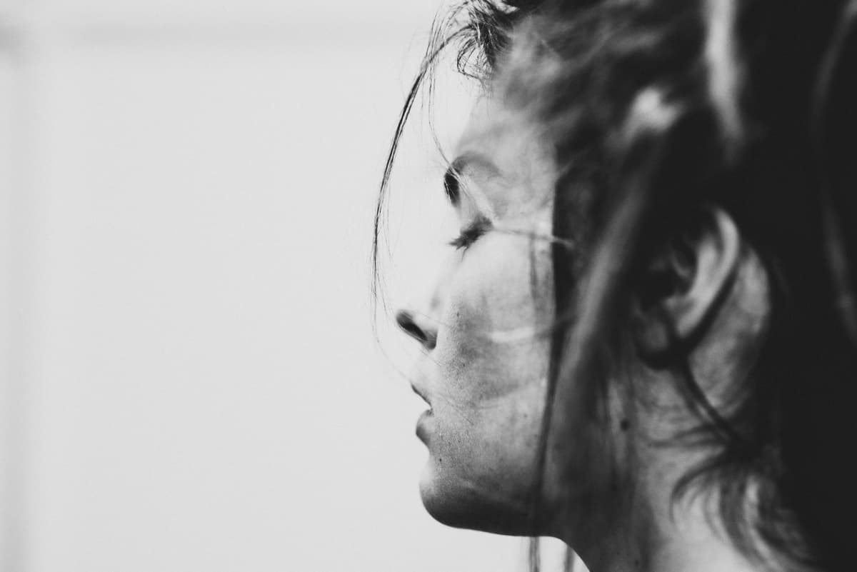 A black-and-white, close-up profile shot of a face, lit from the front with soft light. The eyes are closed, and loose strands of hair hang across the forehead and cheek.