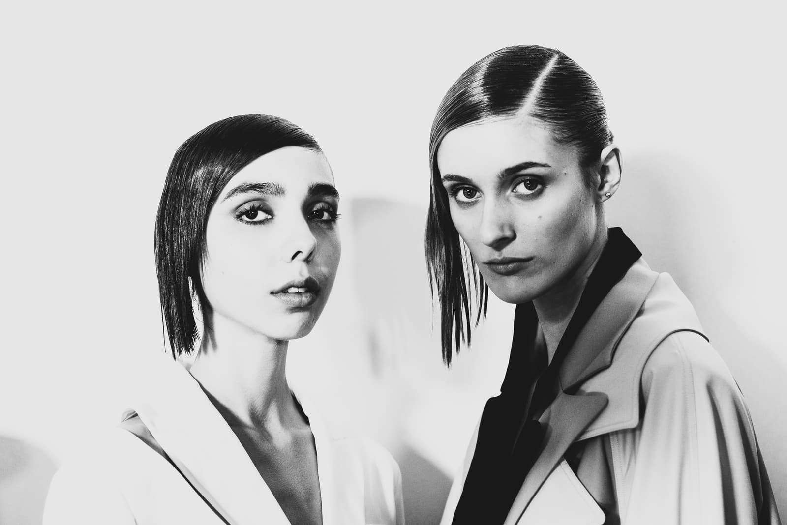 Two people are captured from the chest up against a plain white background. The person on the left has short, sleek dark hair parted deeply to the side, wearing a white garment with a sharp collar. The person on the right has long dark hair styled in a sleek, side-parted wet look, wearing a structured light-coloured trench coat over a dark shirt. Both subjects are looking directly at the camera with neutral expressions in high-contrast black-and-white lighting.