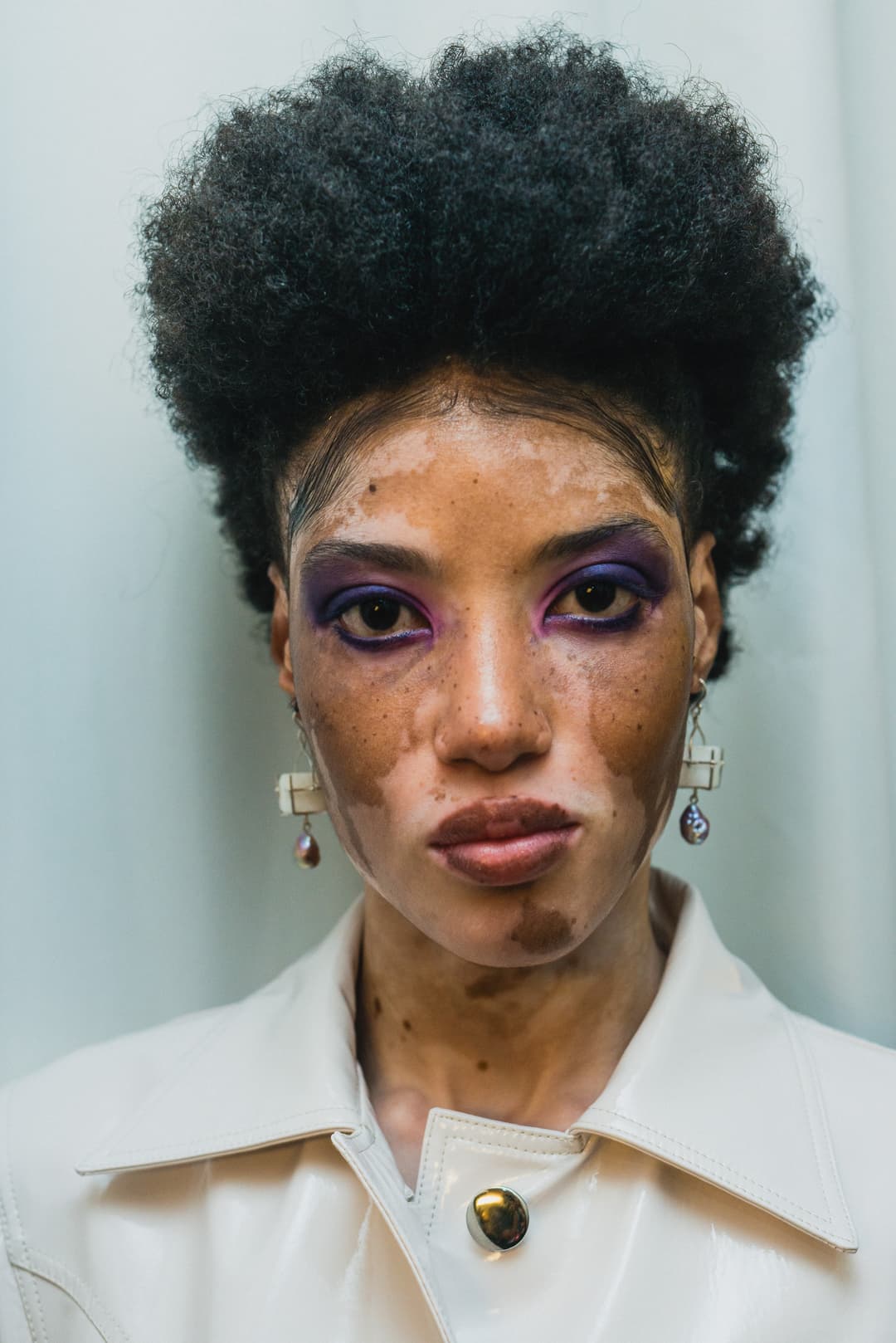 A close-up portrait of a person with dark, textured hair styled upward, wearing bright purple eyeshadow and silver dangling earrings. The skin features visible pigmentation variations and a neutral expression, set against a plain, light-colored background. The person wears a pale, smooth, structured collared garment.