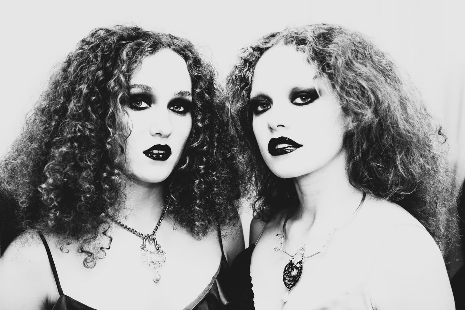 Close-up of two people side by side, both with voluminous curly hair and dark, dramatic eye and lip makeup. Each wears a delicate chain necklace featuring a decorative pendant, against a neutral white background, captured in high-contrast black and white.