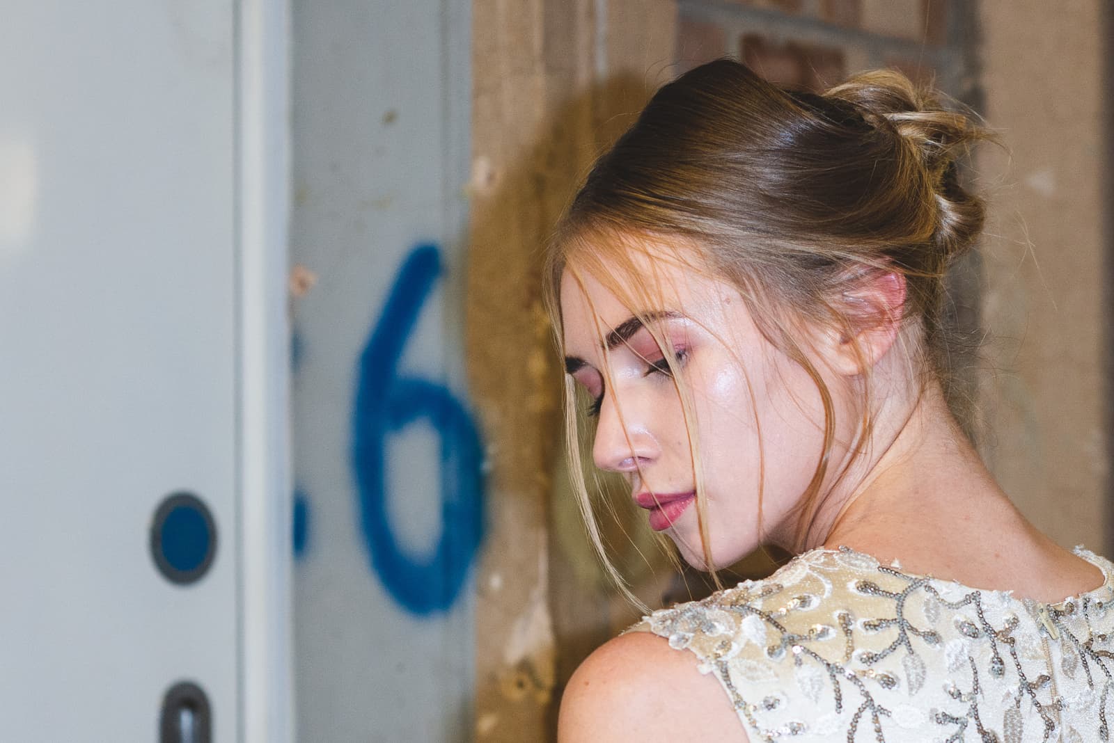 A close-up, over-the-shoulder shot of a person with their head turned away from the camera, wearing an off-white sleeveless garment heavily embellished with silver sequins and intricate beadwork. The person has their hair pulled back in a loose bun with loose strands framing their face, which is visible in profile. The setting is a backstage corridor with a white door featuring a painted blue number, standing against a rough, light-coloured wall.