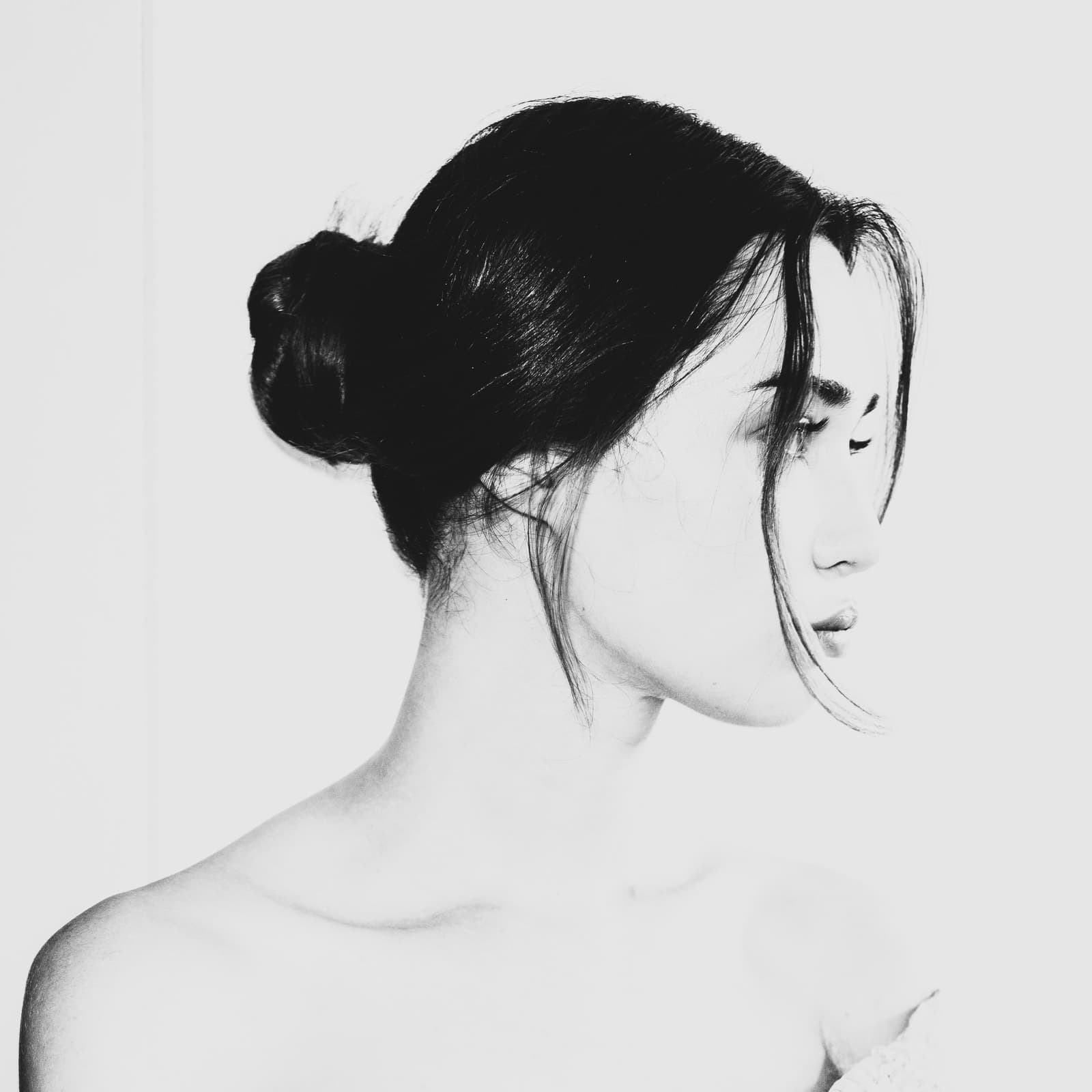 A close-up black and white portrait of a person in profile facing right, with dark hair pulled back into a bun and two loose strands framing the face, set against a plain light-colored background.