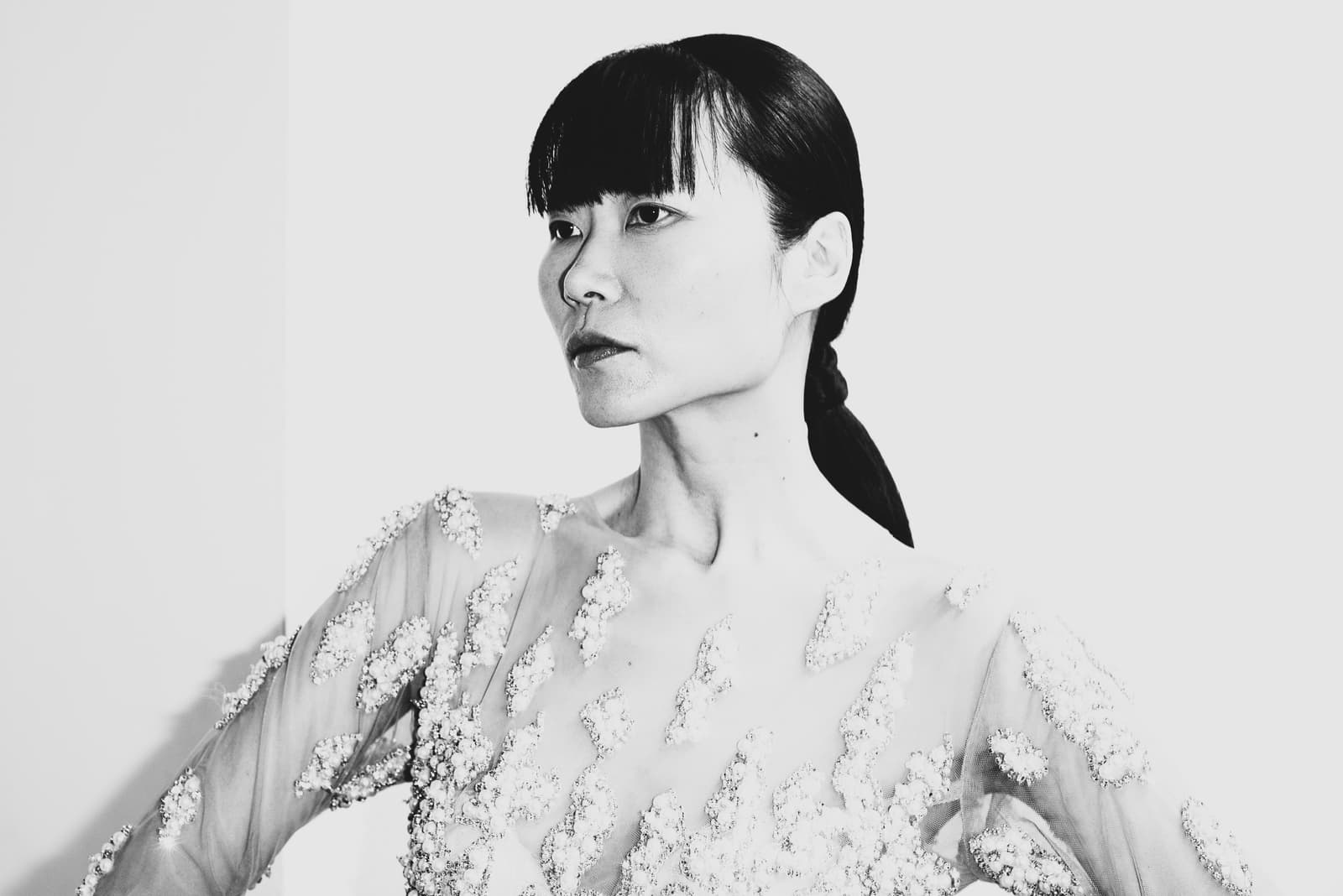 A medium shot in black and white of a person with dark hair styled in a ponytail with straight-cut bangs, posing against a white background. They wear a sheer, long-sleeved top heavily embellished with intricate, leaf-shaped beadwork or embroidery. The person is looking slightly off-camera to the left with a neutral expression.