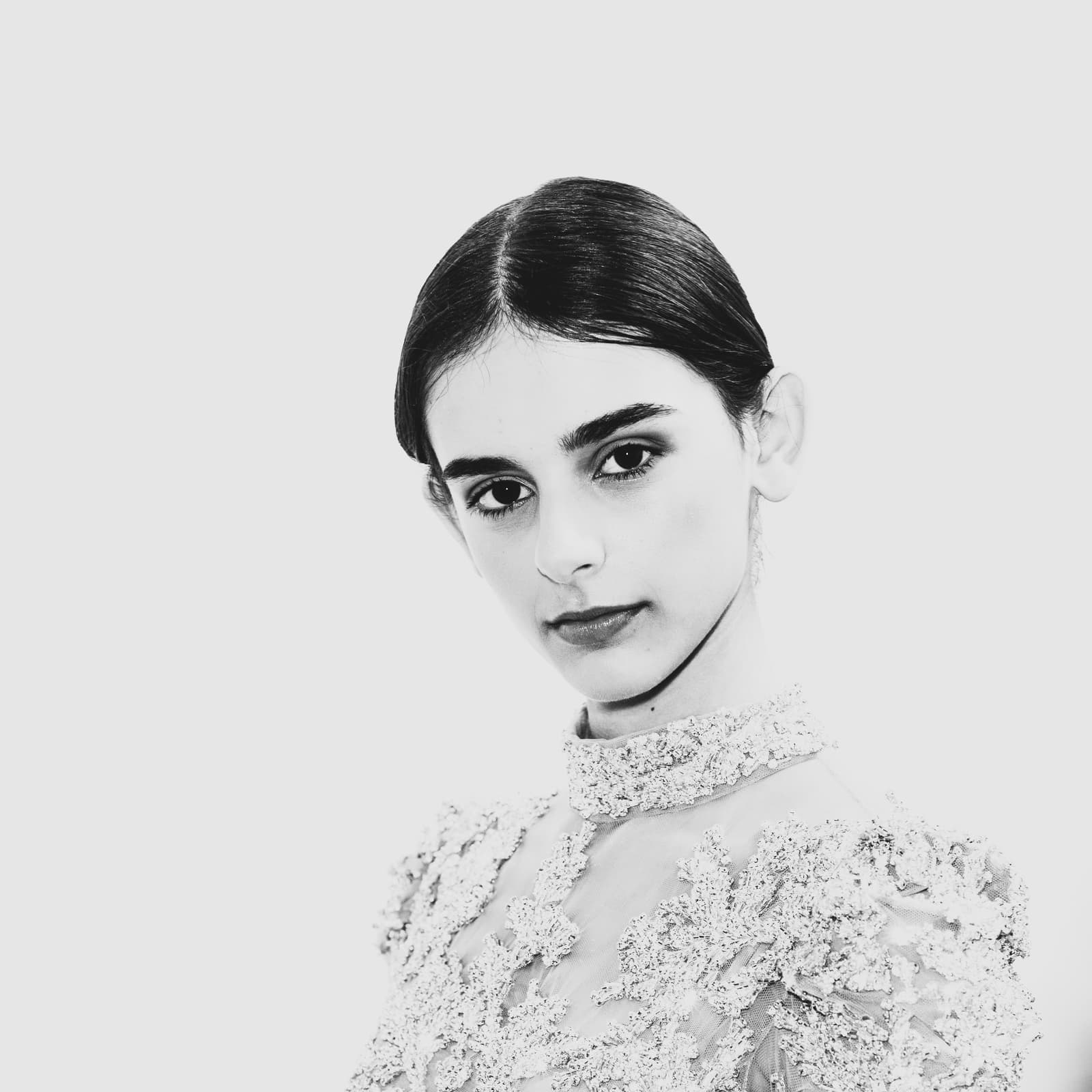 A close-up black and white portrait of a person with dark hair pulled back in a neat center part. They are wearing a high-necked garment with intricate, heavy lace or floral embroidery across the chest and shoulders, against a plain, bright white background.
