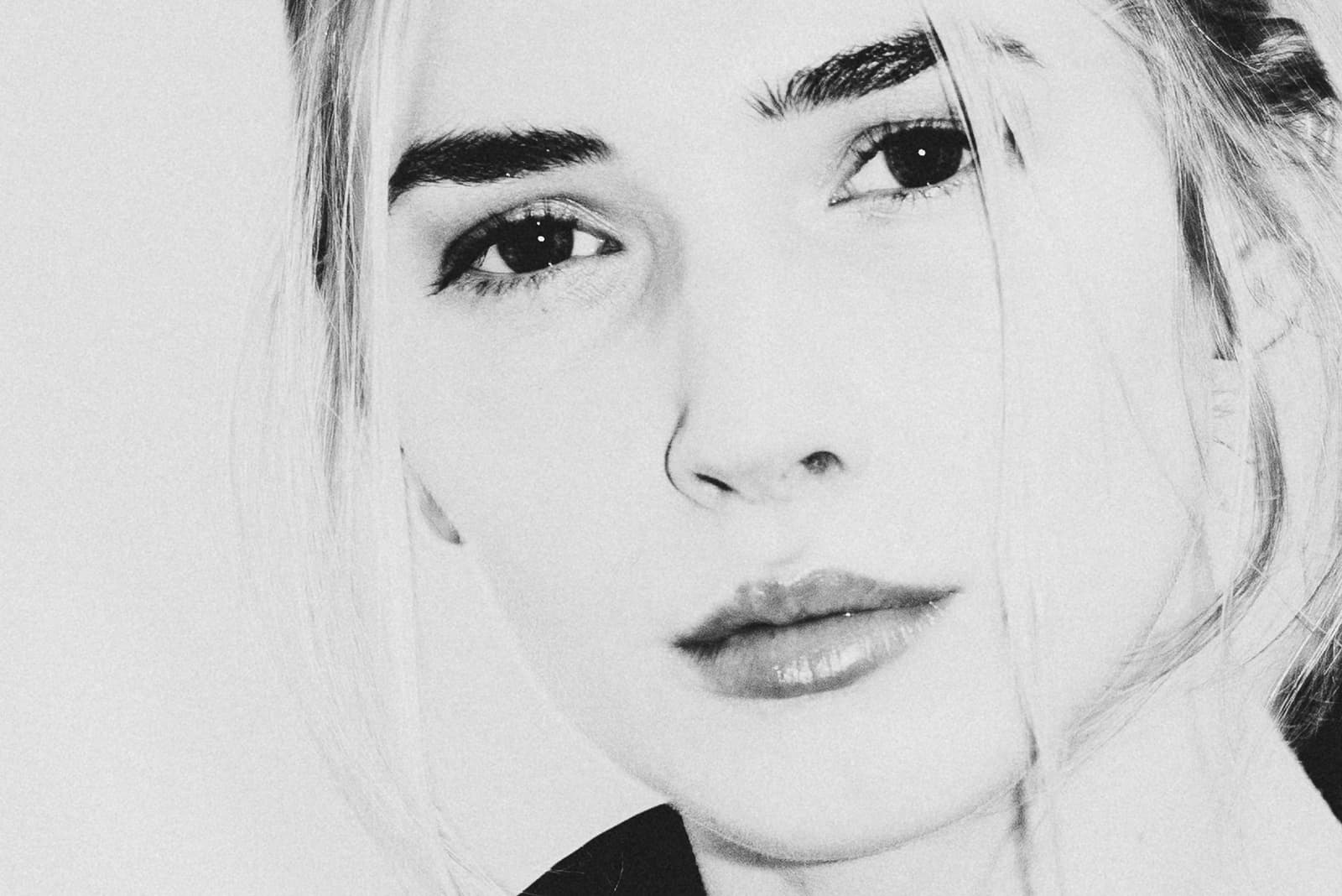 A tight, high-key close-up portrait of a face, lit with bright, flat light against a white background. Dark eyebrows and eyeliner contrast with very light, blonde hair framing the face. The expression is calm and neutral, with the gaze directed forward.