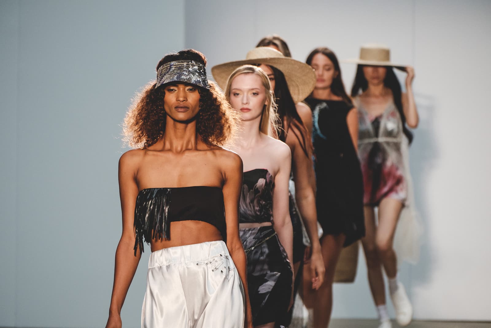 A group of models walking in a single file line down a brightly lit white runway. The model in the foreground wears a black strapless top with fringe detail, a sparkling silver visor, and white trousers. Behind her, other models walk in dresses and summer attire, with some wearing wide-brimmed straw hats.