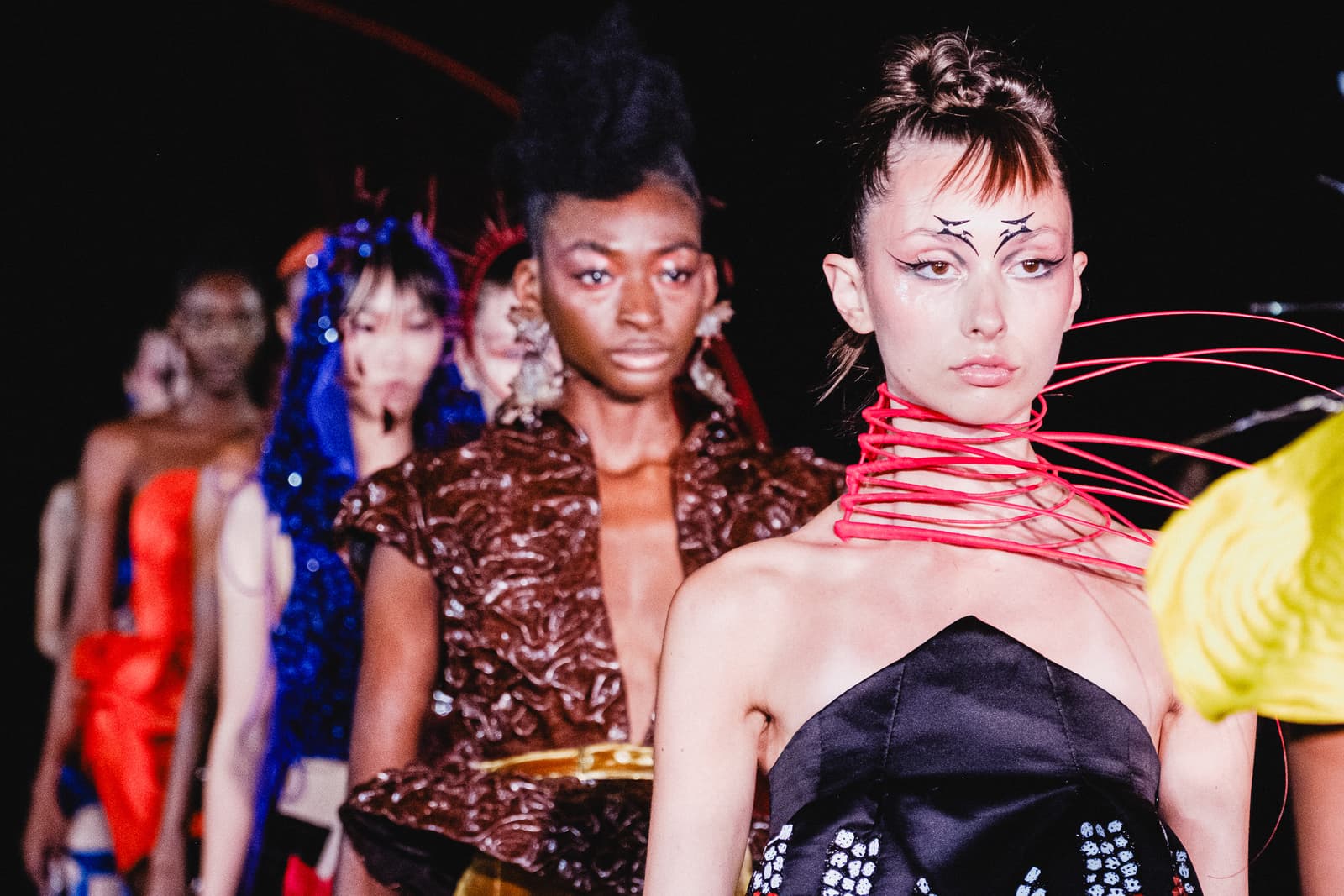 A line of fashion models walking down a runway in a dark space. The focus is on a model in the foreground wearing a black strapless garment, with bright red wire-like structures coiled around her neck and chest. She has distinctive eye makeup with sharp lines extending above the brow. Behind her, other models in elaborate textured and colourful clothing follow in a procession, illuminated by stage lighting against a black background.