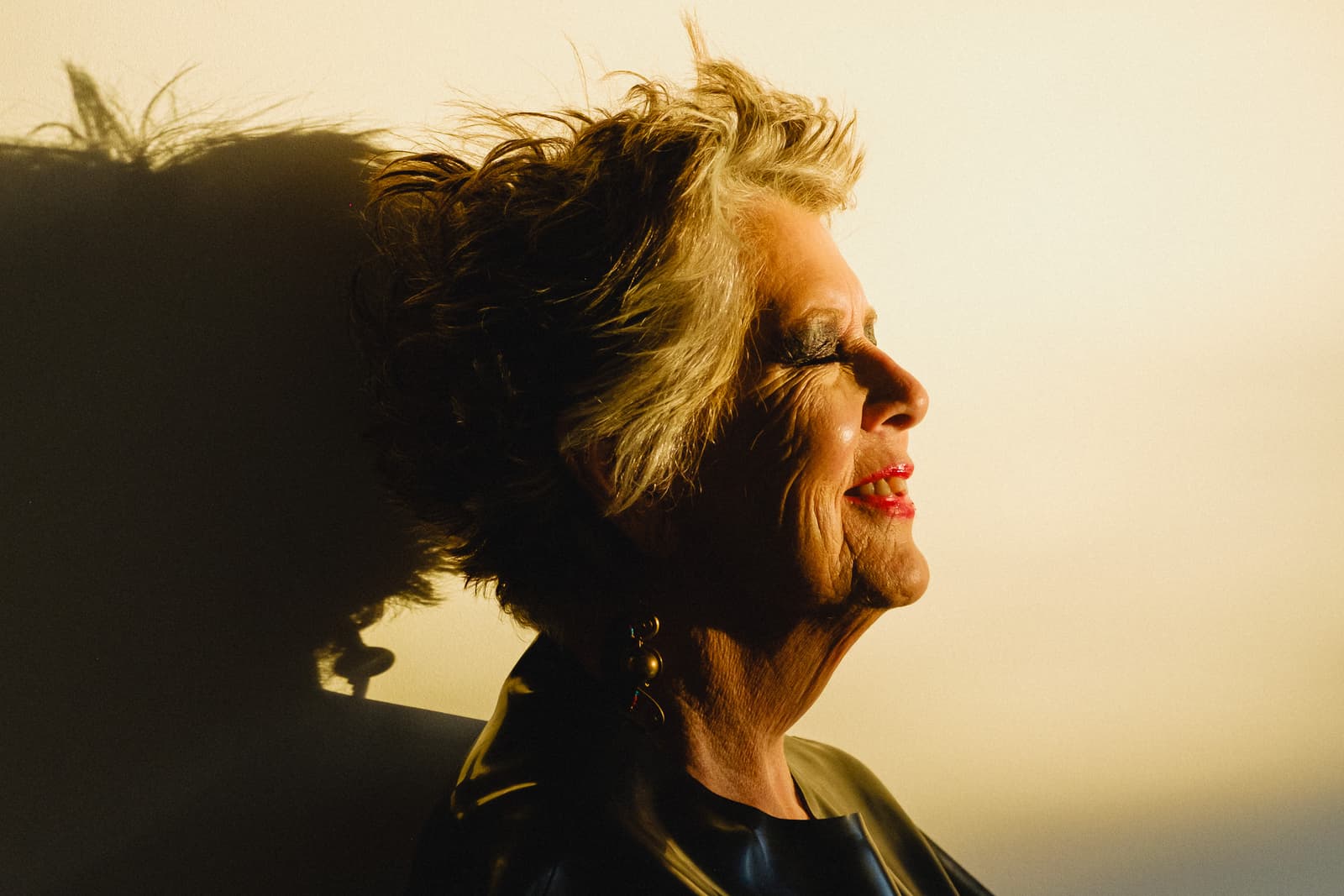 A close-up profile view of a person with short, textured grey hair against a plain, light-toned background. Harsh, warm-toned light casts a distinct, dark shadow of the head and shoulders onto the surface behind them. The person is wearing dark clothing and simple, dangling drop earrings. The light catches the texture of their skin and highlights the side of their face.
