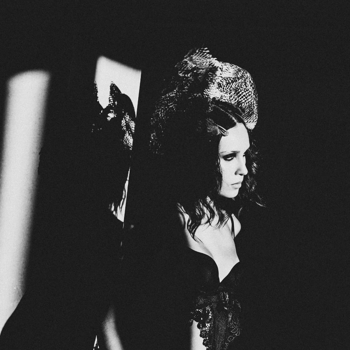 A black-and-white medium shot of a person facing right, wearing an ornate, textured headpiece and a dark, lace-embellished garment. Strong light from the left creates a sharp, bright pillar of illumination, casting a distinct shadow of the person’s profile and headgear onto the background wall.