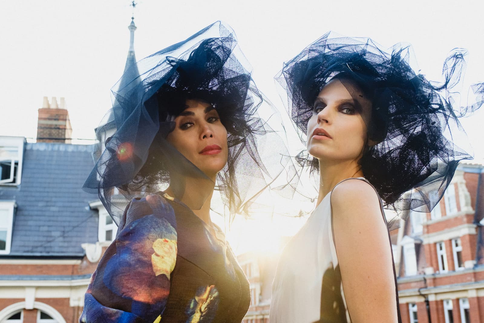 Two people wearing large black tulle fascinators against a bright outdoor background featuring a blurred brick building with windows. Both are looking toward the camera; one wears a dark patterned dress while the other wears a simple white sleeveless top, illuminated by backlighting that creates a lens flare.