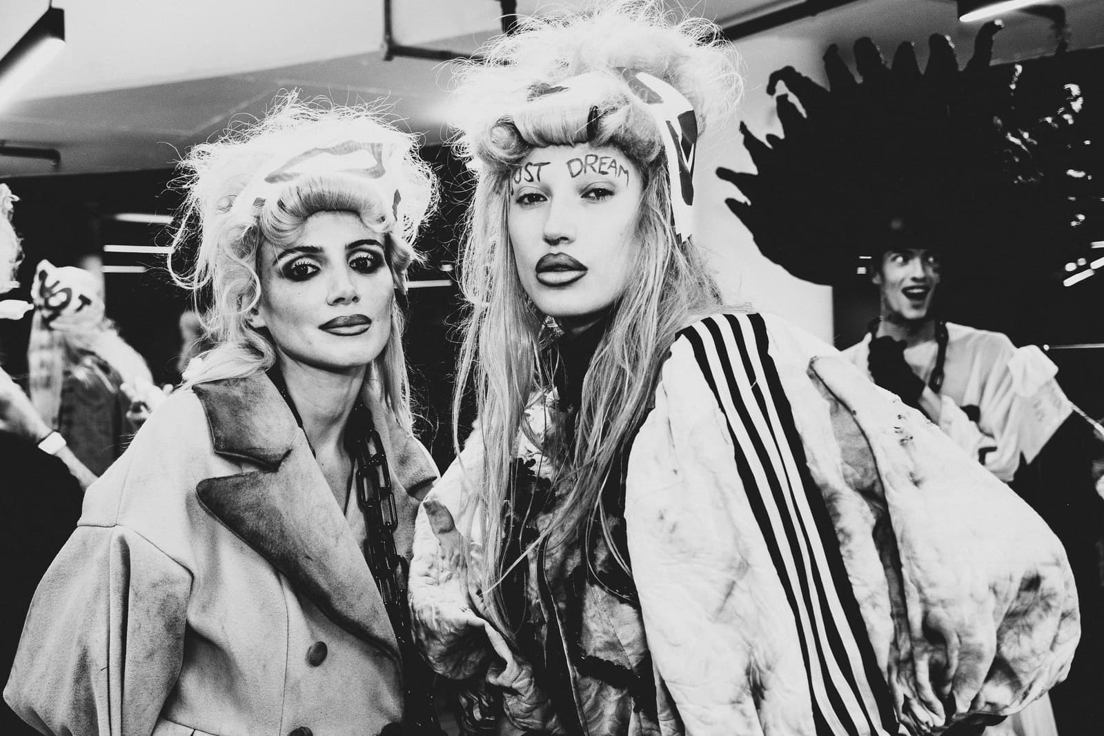 A black-and-white, medium close-up shot of two models backstage at a fashion show. Both have elaborate, blonde, teased-up hairstyles with graphic accessories, and dark, dramatic makeup. The person on the left wears a structured, heavy coat. The person on the right, centrally placed, wears an oversized, athletic-style white jacket with black stripes, and has the words 'JUST DREAM' written in dark ink across their forehead. In the background, out of focus, another person smiles at the camera.