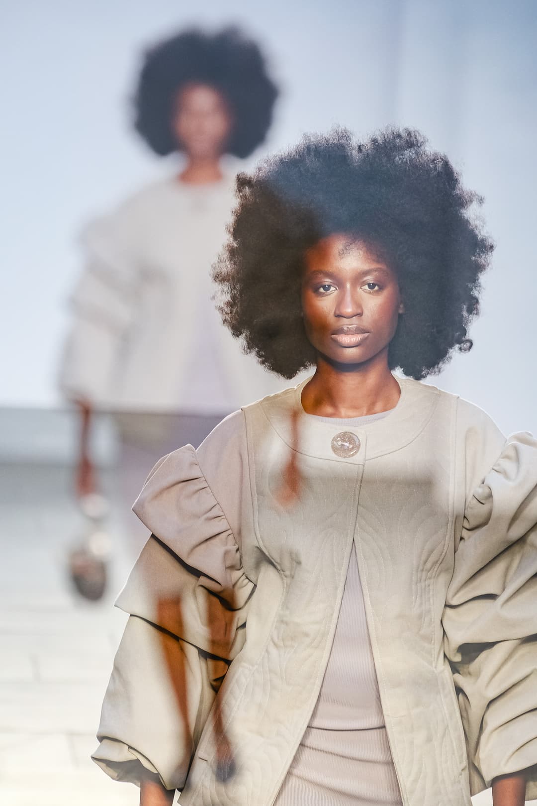 A medium shot of a person with dark skin and a large afro hairstyle walking on a runway. They are wearing a structured, beige quilted jacket with exaggerated, voluminous puffed sleeves. The background is a soft, out-of-focus white, with another person slightly blurred in the distance. A decorative, dark textured element appears as a blurred overlay in the foreground.
