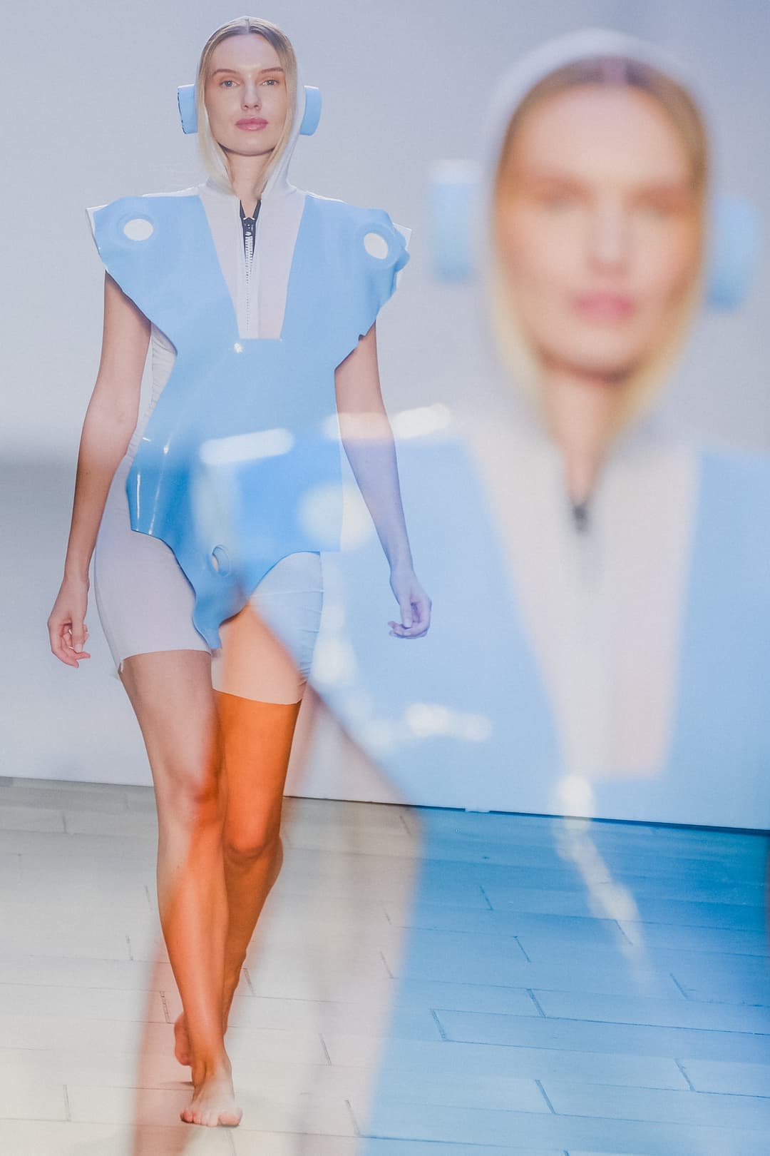 A model with blonde hair walks barefoot down a catwalk, wearing a pale blue structured tabard with circular cutouts over a cream-colored zippered hooded bodysuit. In the foreground, an out-of-focus, semi-transparent reflection of the same person is superimposed over the right side of the frame.