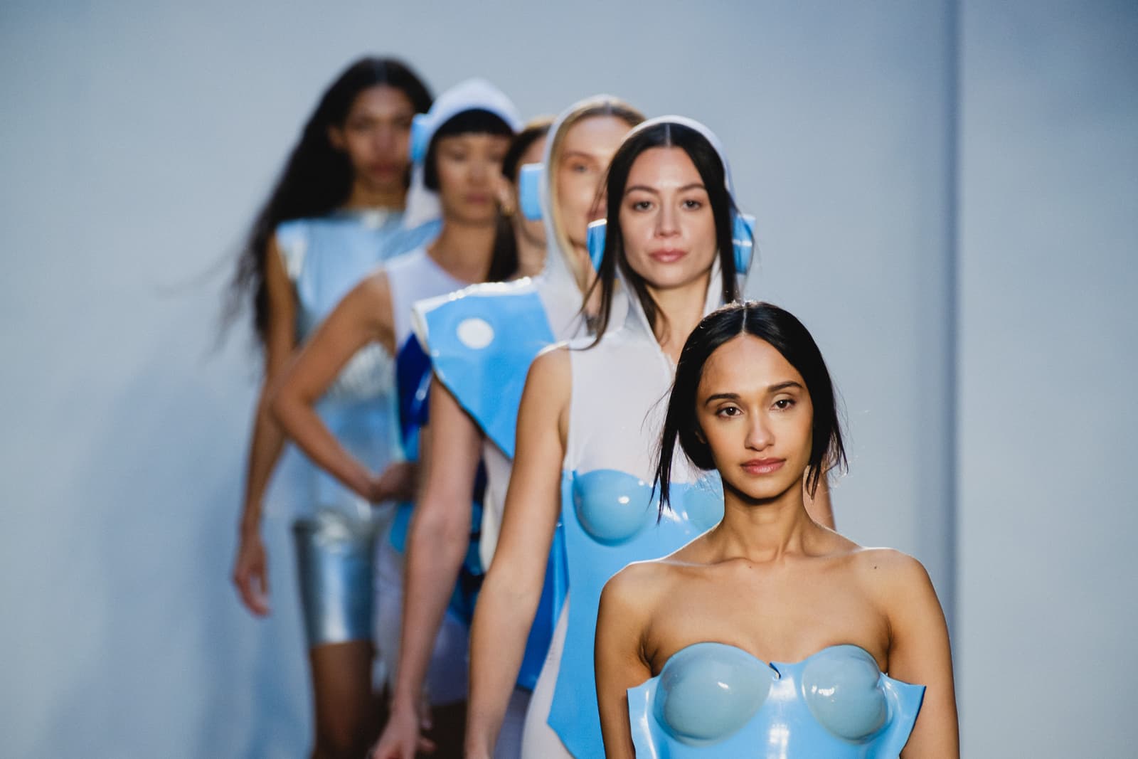 A line of models walking in a fashion show, seen in a shallow depth of field shot. The foreground model wears a strapless, light blue molded plastic top, while the models behind her wear white and blue structured garments, some with hoods or headpieces, all against a plain light grey backdrop.