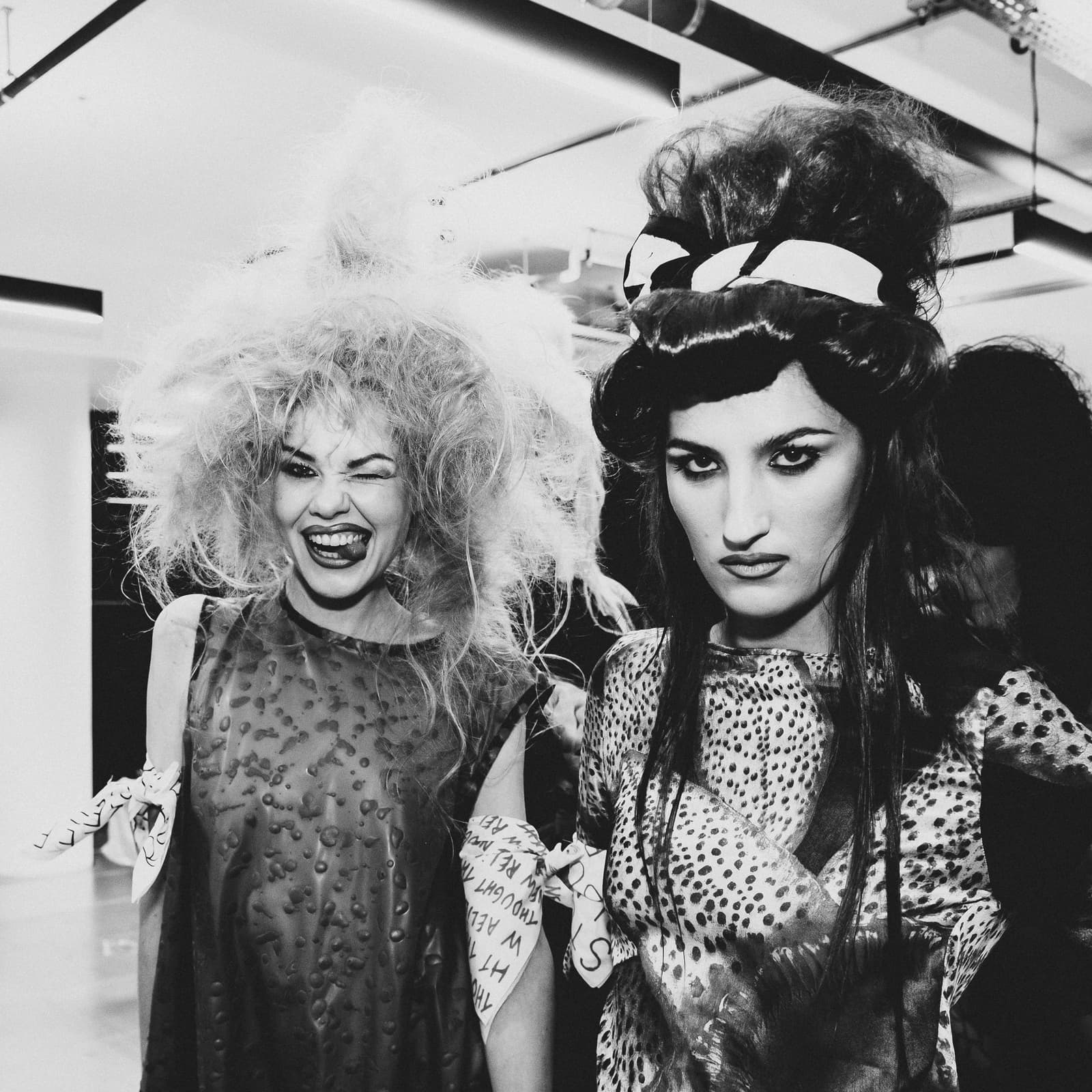 Two models standing side-by-side backstage. The model on the left has large, teased, blonde hair and is winking and sticking out her tongue, wearing a textured sleeveless top. The model on the right has dark, voluminous, styled hair tied with a patterned ribbon, wearing a printed top, looking directly at the camera with a neutral expression.