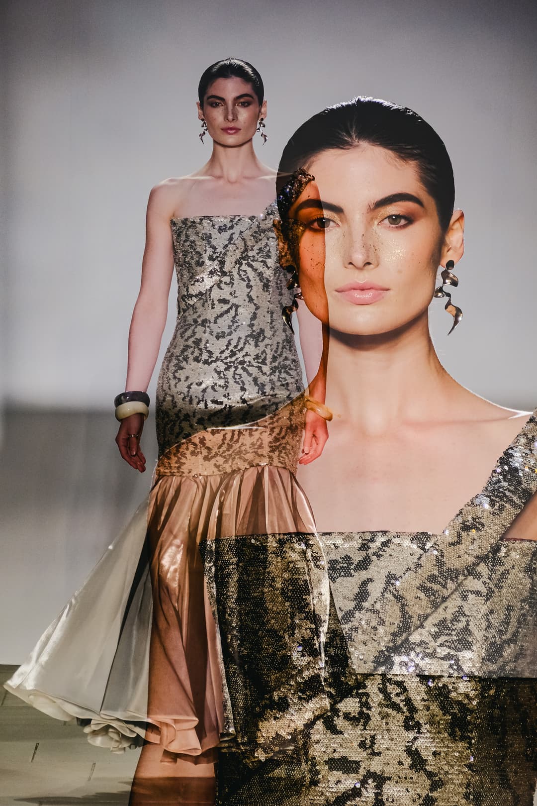 A double exposure photograph of a fashion model on a runway. In the background layer, the model walks in a strapless, floor-length gown featuring metallic, sequined textured fabric and a flared hem. In the foreground layer, a large, translucent close-up portrait of the model's face shows bronzed makeup, slicked-back hair, and sculptural silver earrings, with fine glitter flecks visible across the skin.