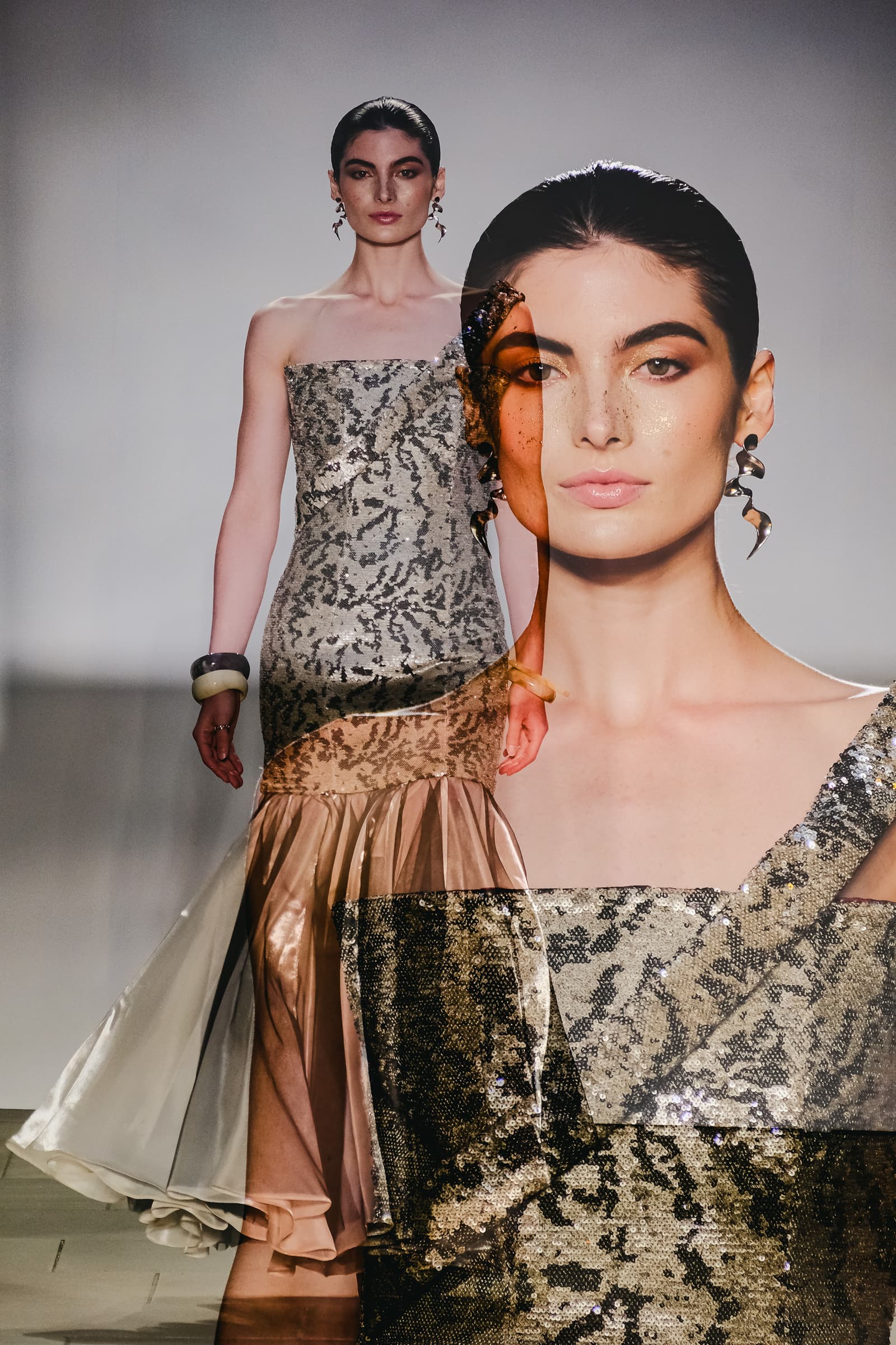 A double exposure photograph of a fashion model on a runway. In the background layer, the model walks in a strapless, floor-length gown featuring metallic, sequined textured fabric and a flared hem. In the foreground layer, a large, translucent close-up portrait of the model's face shows bronzed makeup, slicked-back hair, and sculptural silver earrings, with fine glitter flecks visible across the skin.