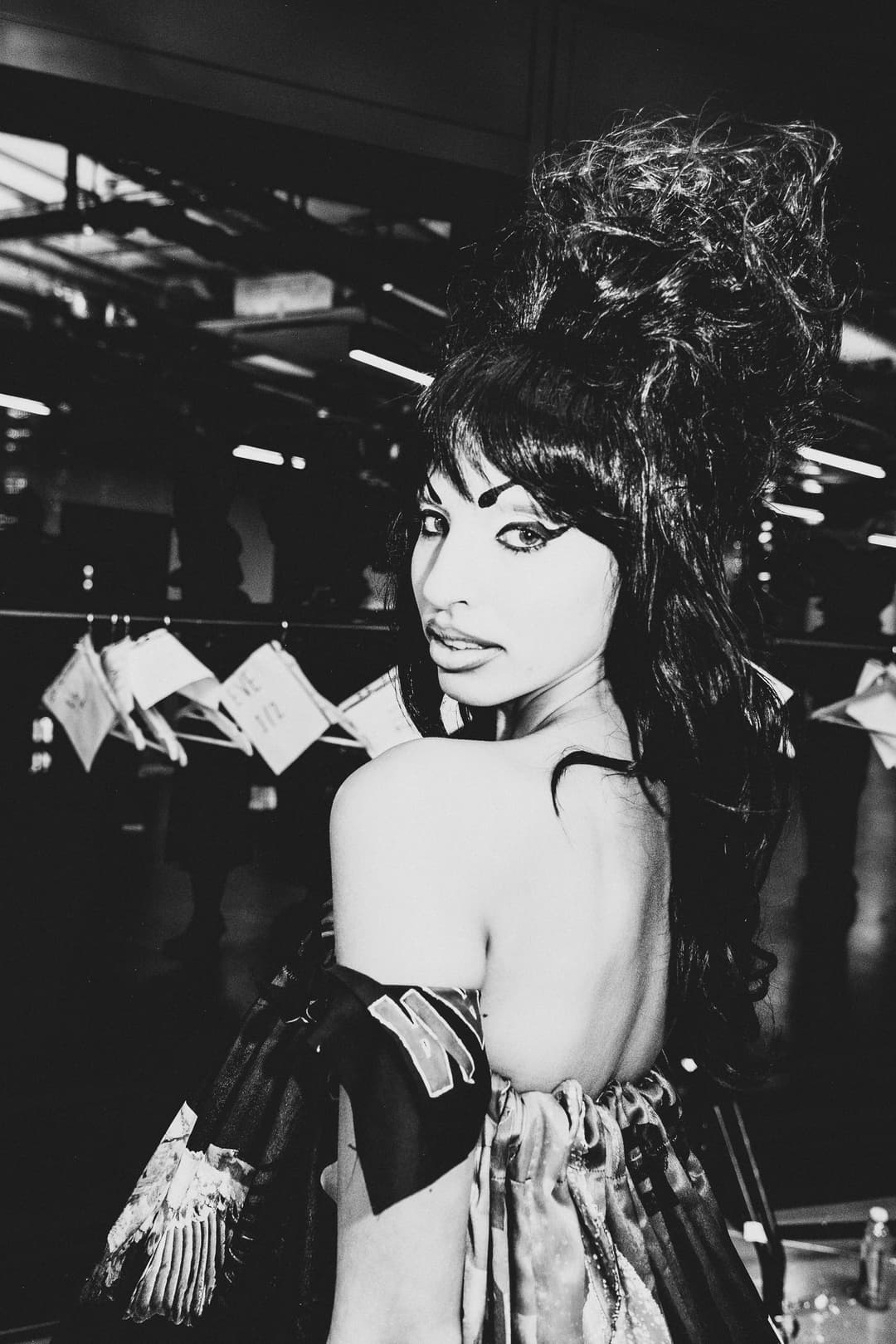 A black-and-white medium shot taken backstage at a fashion show. A person with tall, bouffant-style dark hair and winged eyeliner looks over their shoulder toward the camera. They wear an off-the-shoulder garment with visible textures and patterns. In the blurred background, clothing racks with white tags are visible in the dimly lit space.