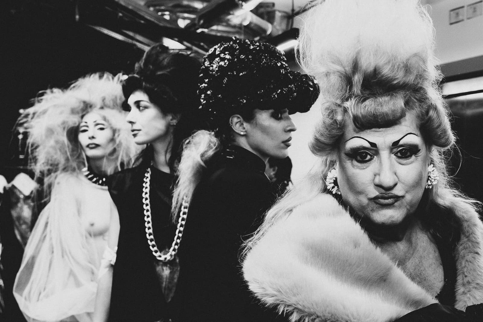 A black-and-white backstage photograph featuring four individuals with elaborate hair and makeup at a fashion show. In the foreground right, a person with highly styled, voluminous hair and dramatic eyeliner looks directly at the camera wearing a fur stole. To the left, three other figures are positioned in profile or three-quarter view, wearing dark, textured clothing, large chain necklaces, and varied structured headpieces.