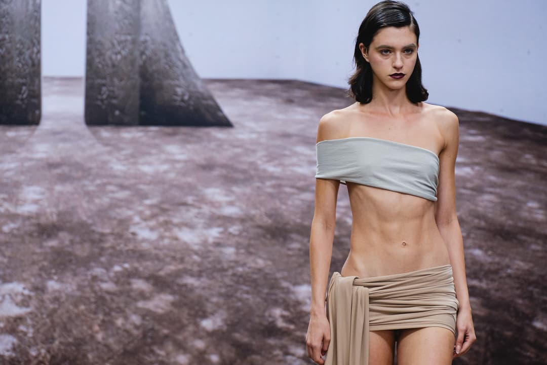 A model walks on a dark, mottled-pattern runway with large sculptural geometric props in the background. The model wears a light sage-green bandeau top and a beige wrap-around skirt. The model has dark, shoulder-length hair and dark-coloured lipstick.