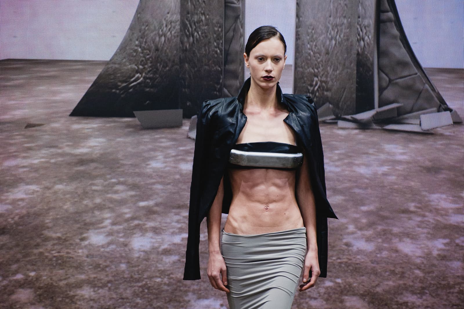 A model walks on a catwalk wearing a black leather jacket draped over the shoulders, a silver-toned bandeau top across the midriff, and a light gray skirt. The background features large, abstract, metallic-textured geometric structures on a muted, brownish ground. The model has dark lipstick and hair pulled back, with a focused expression.