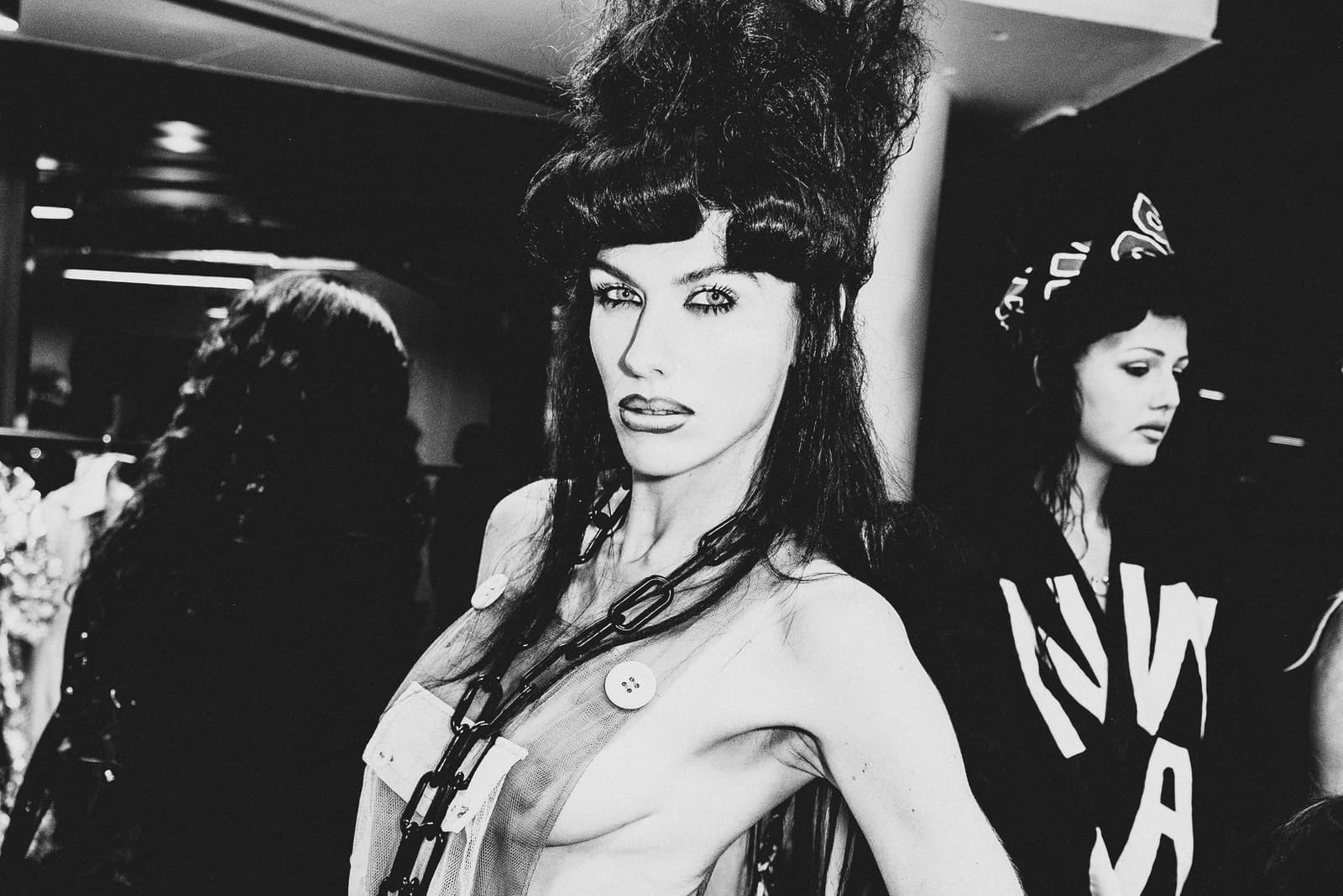 A black-and-white medium shot of a model backstage at a fashion show. The subject wears a sheer, sleeveless garment with a prominent black plastic chain draped across the chest and large buttons attached. Her hair is styled in an oversized, voluminous bouffant. She is looking directly at the camera with a steady expression, while another individual in a black hoodie with graphic white lettering stands partially out of focus to the right.