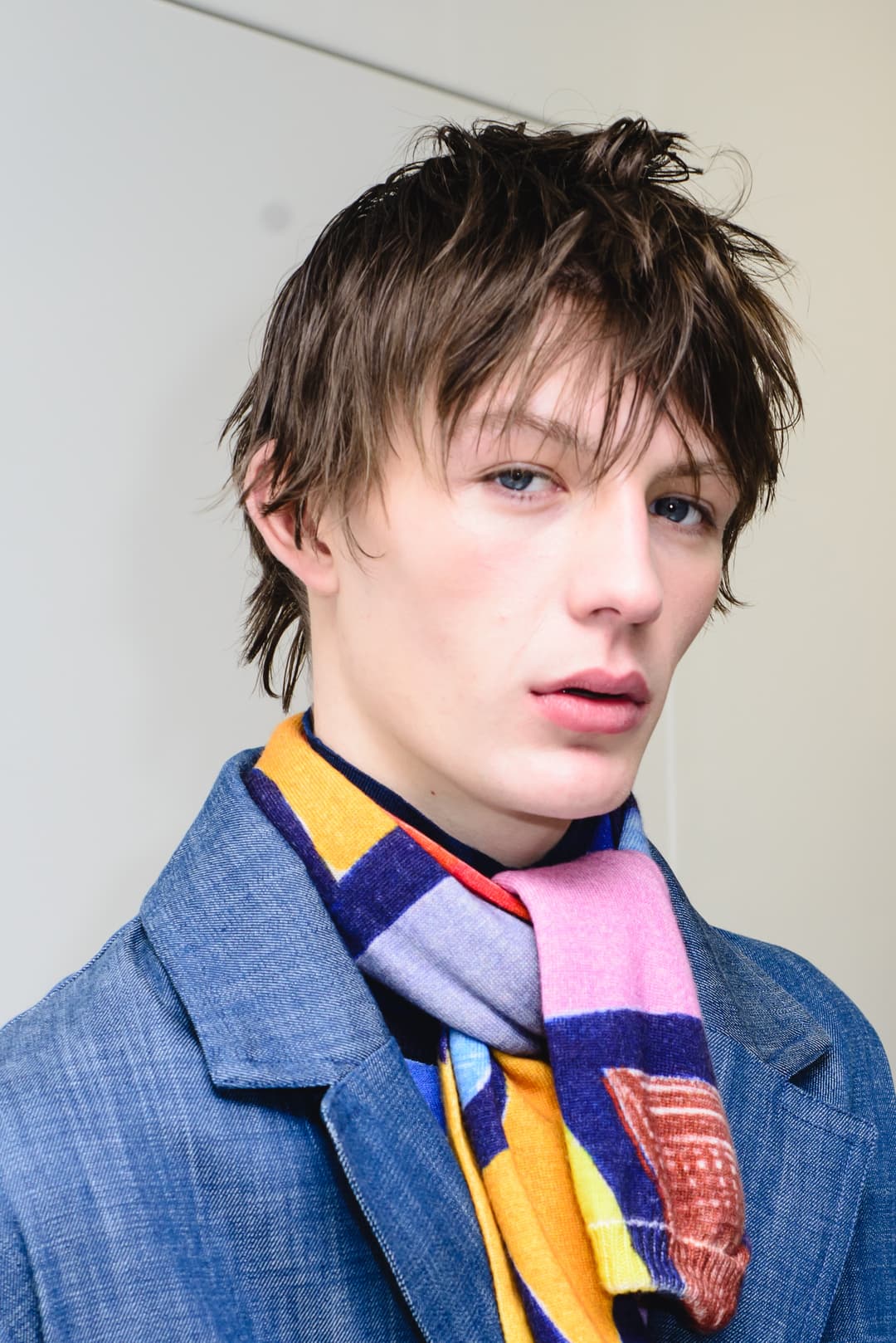 A close-up portrait of a person with textured brown hair looking directly into the camera. They are wearing a blue denim jacket layered over a dark sweater and a bright, multi-coloured geometric knit scarf. The background is a simple, off-white wall.