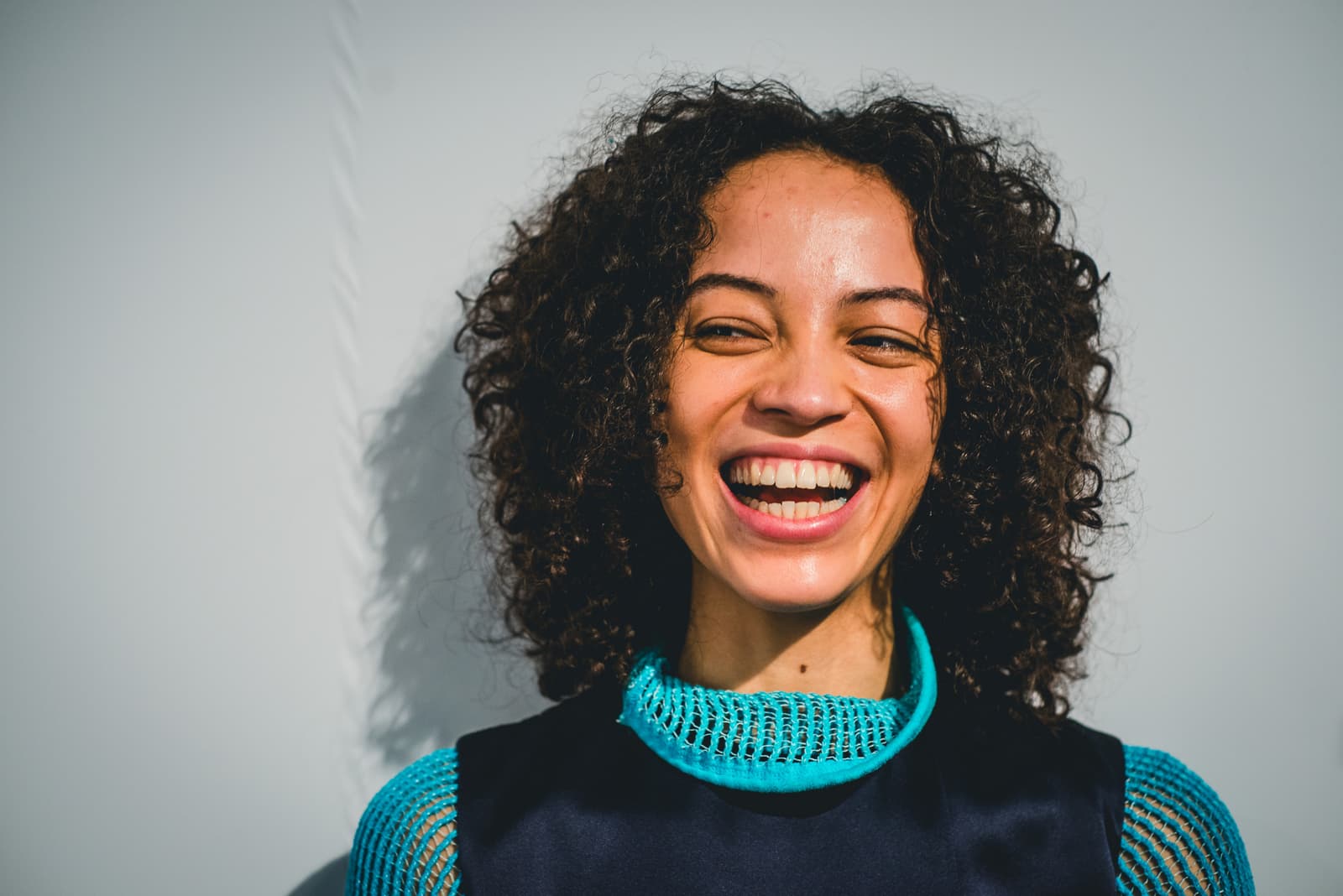 A medium shot of a person with dark curly hair laughing, captured in bright, direct sunlight against a pale grey wall. The person wears a dark sleeveless top layered over a bright turquoise mesh turtleneck piece that frames the neck.