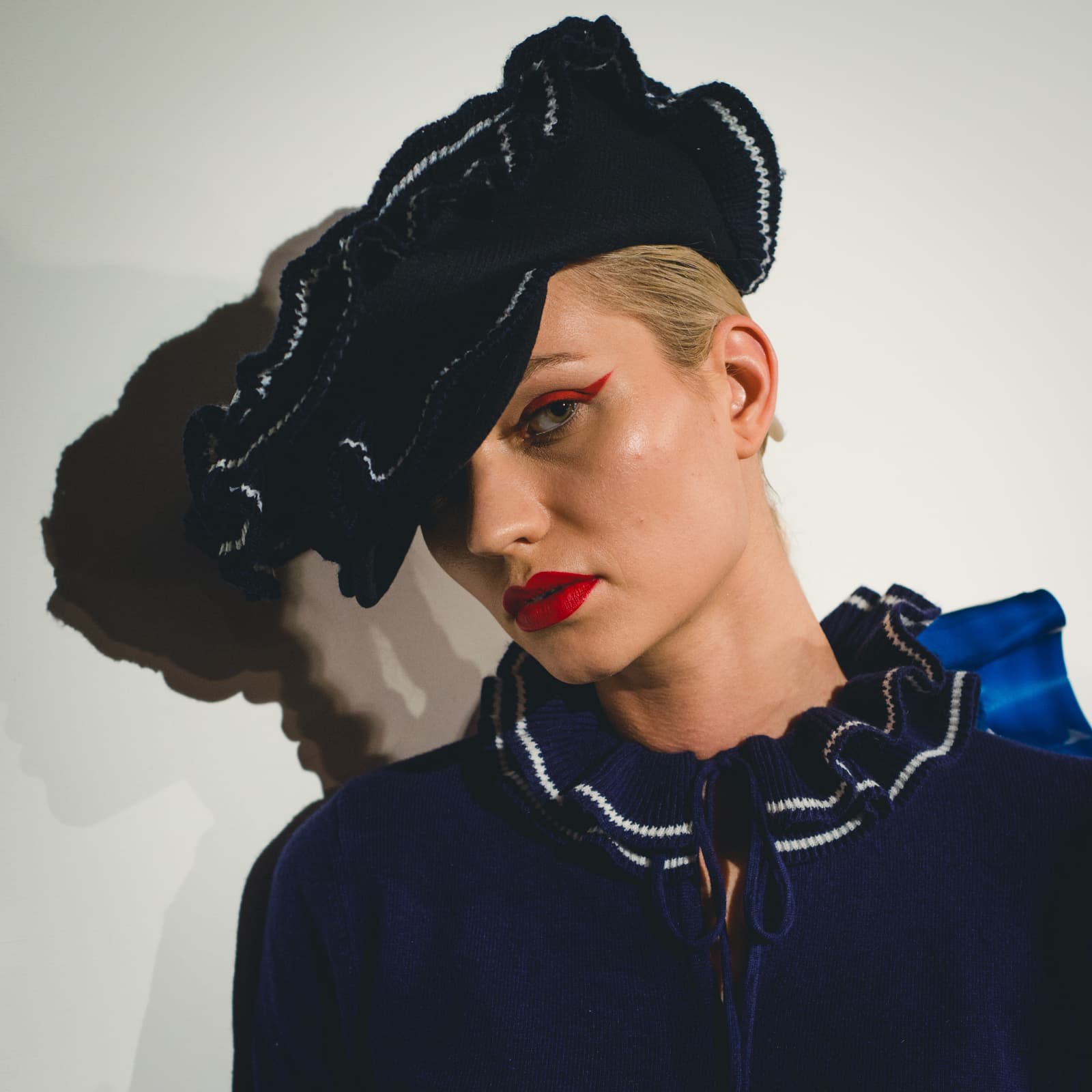 Close-up portrait of a person with blonde hair wearing a dark blue ruffled knit top and a matching sculptural beret with white stitching. The person has bright red winged eyeliner and bold red lipstick, looking directly at the camera. Harsh studio lighting casts a prominent shadow against a white background.