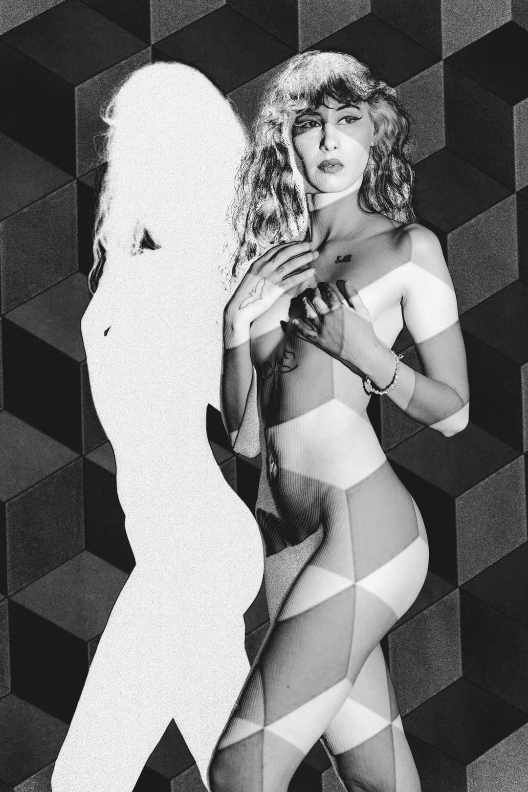 A black and white studio photograph of a person from the chest up, posed against a geometric backdrop. A projection of repeating cubes and angular patterns overlays the subject's skin and the dark background. To the left of the subject, a bright, solid white silhouette of a figure is cast onto the wall, while the subject stands facing forward, hands held near the chest. The image has a solarised, high-contrast aesthetic.