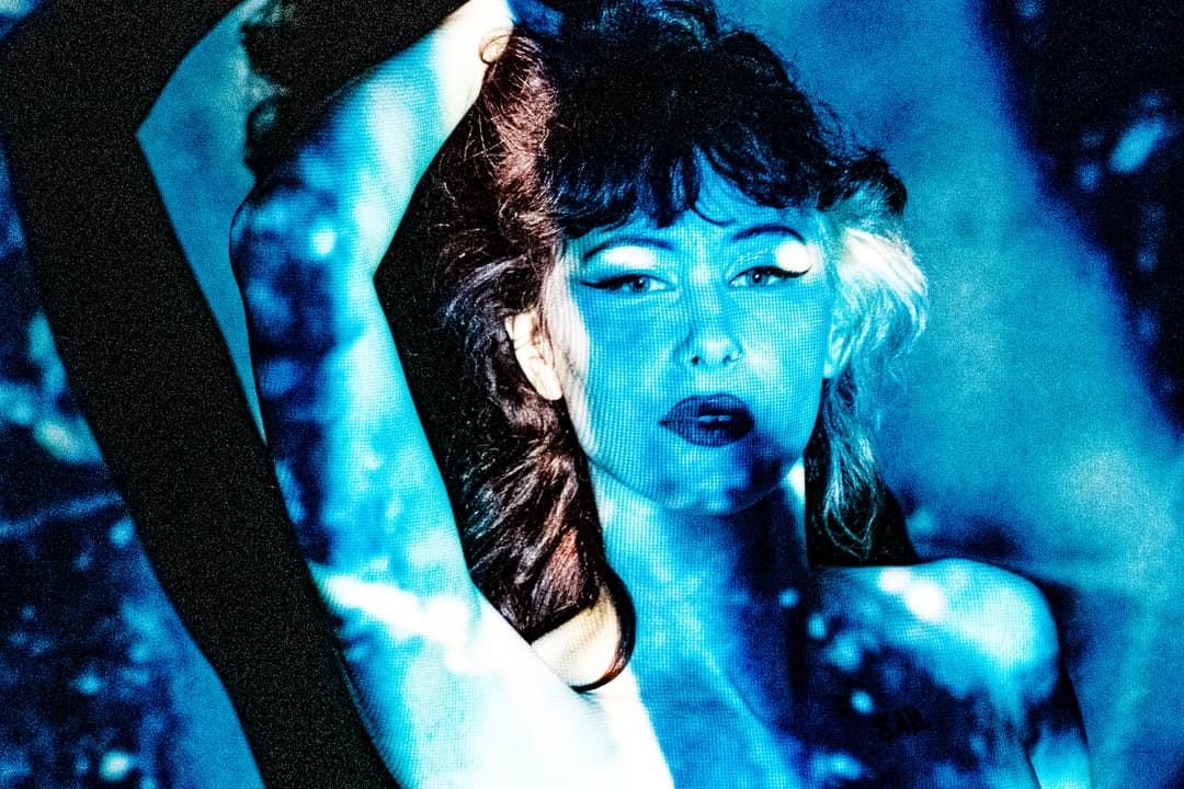 A close-up shot of a person with dark hair and heavy eyeliner, illuminated by a high-contrast blue light projection that casts a grid-like pattern across their skin. One arm is raised above their head. The image is highly grainy with deep blue and black tones.