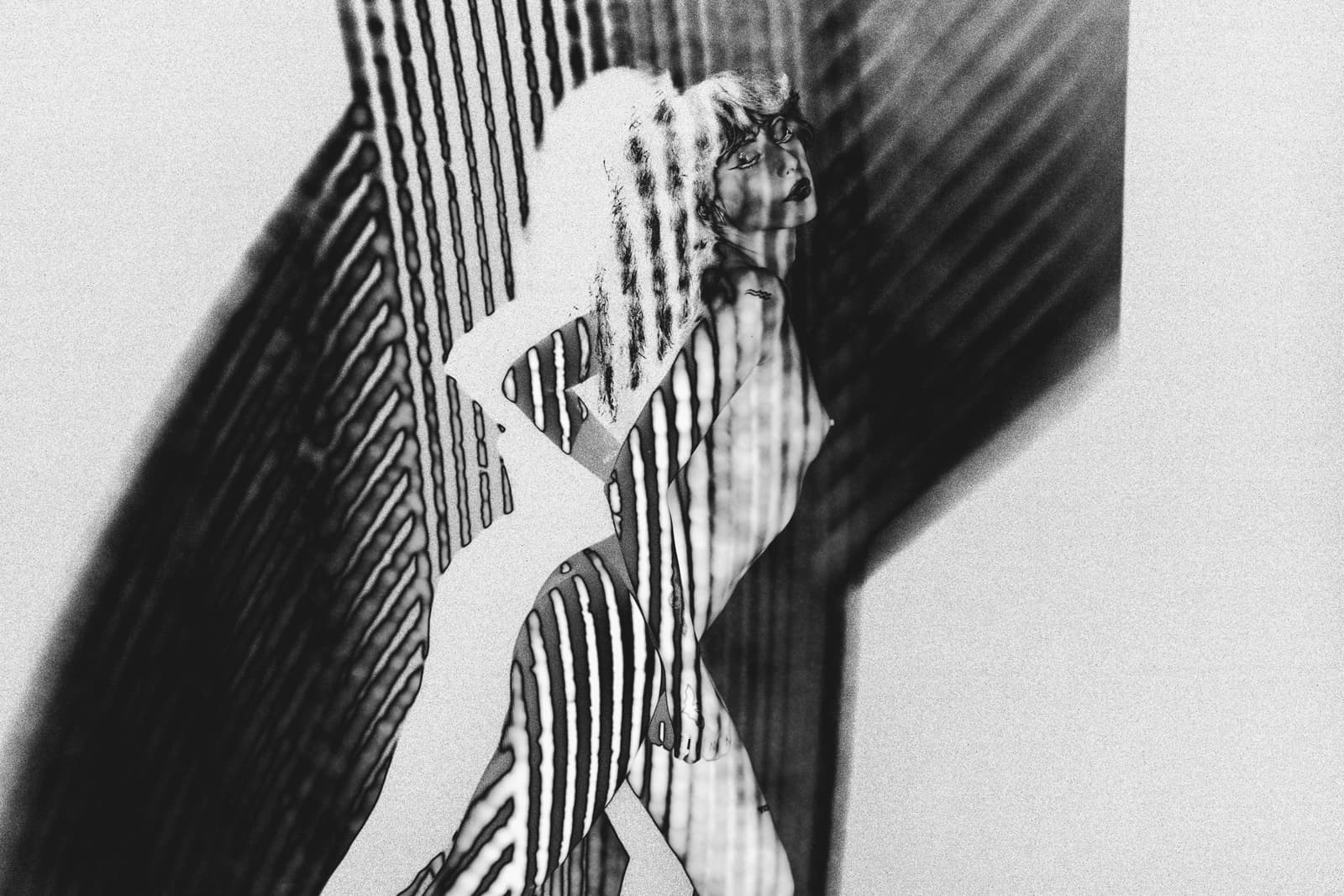 A black and white solarised image of a person seen from the side, standing with one hand on their hip. Patterns of vertical stripes are projected over the body and the background, creating high-contrast graphic lines across the skin and surrounding space.