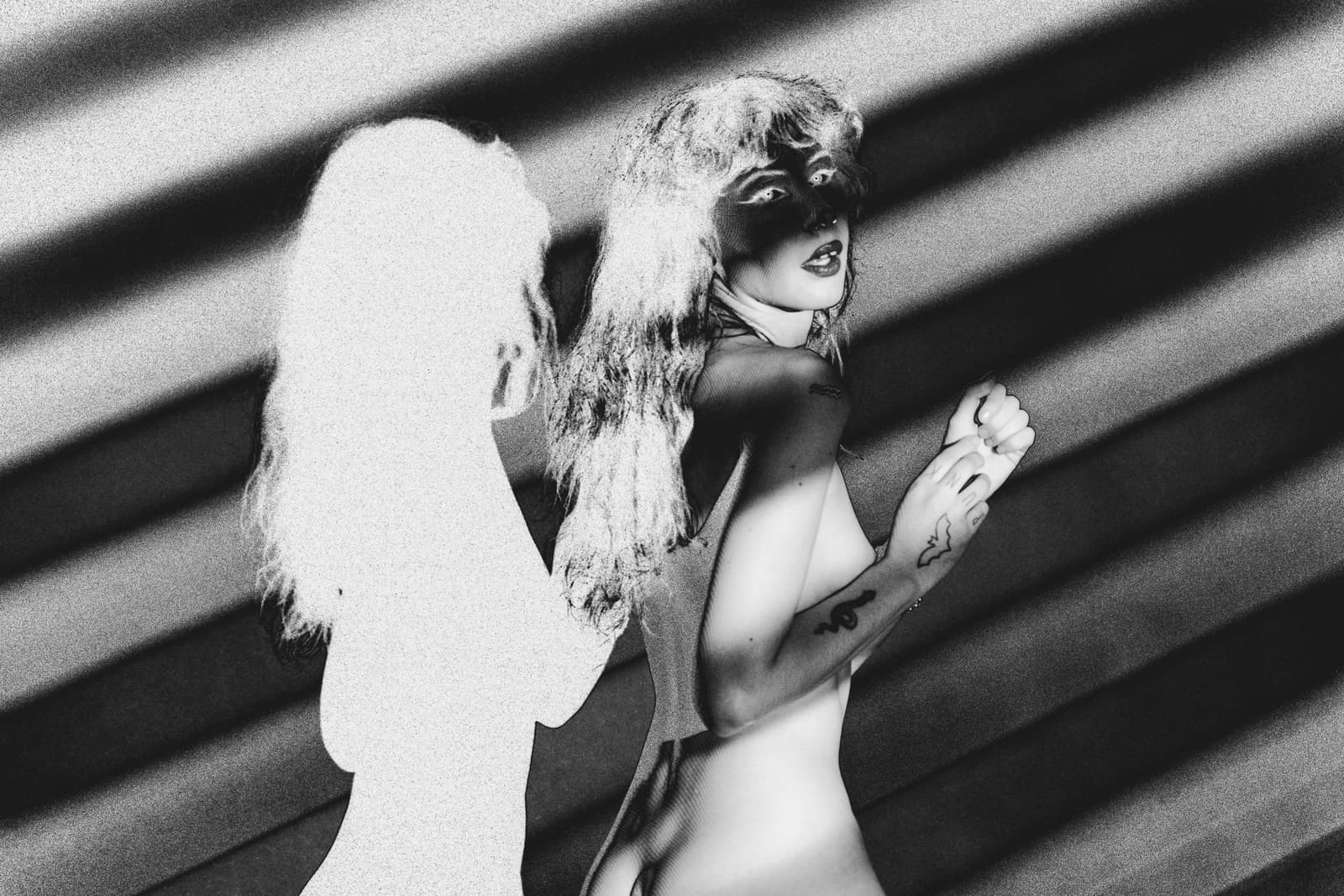A person viewed from the back and side, shown against a background of dark, diagonal lines. A solarised image of another person is projected over them, blending their form with light-coloured silhouettes and harsh, high-contrast shadows. The person's hair is wavy and light, and their hands are raised near their chest.