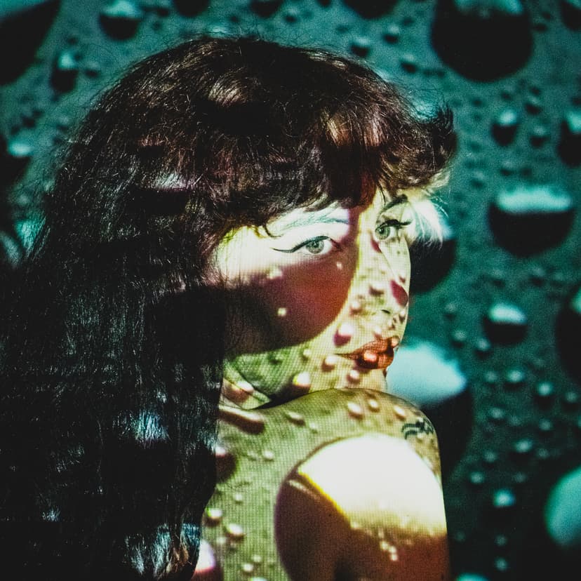 A close-up shot of a person with dark hair looking over their shoulder toward the left edge of the frame. A projection of large, round, translucent water droplets covers the subject's skin and face in soft teal and white light. The background is a dark, textured wall with the same droplet pattern. The subject wears dark winged eyeliner.