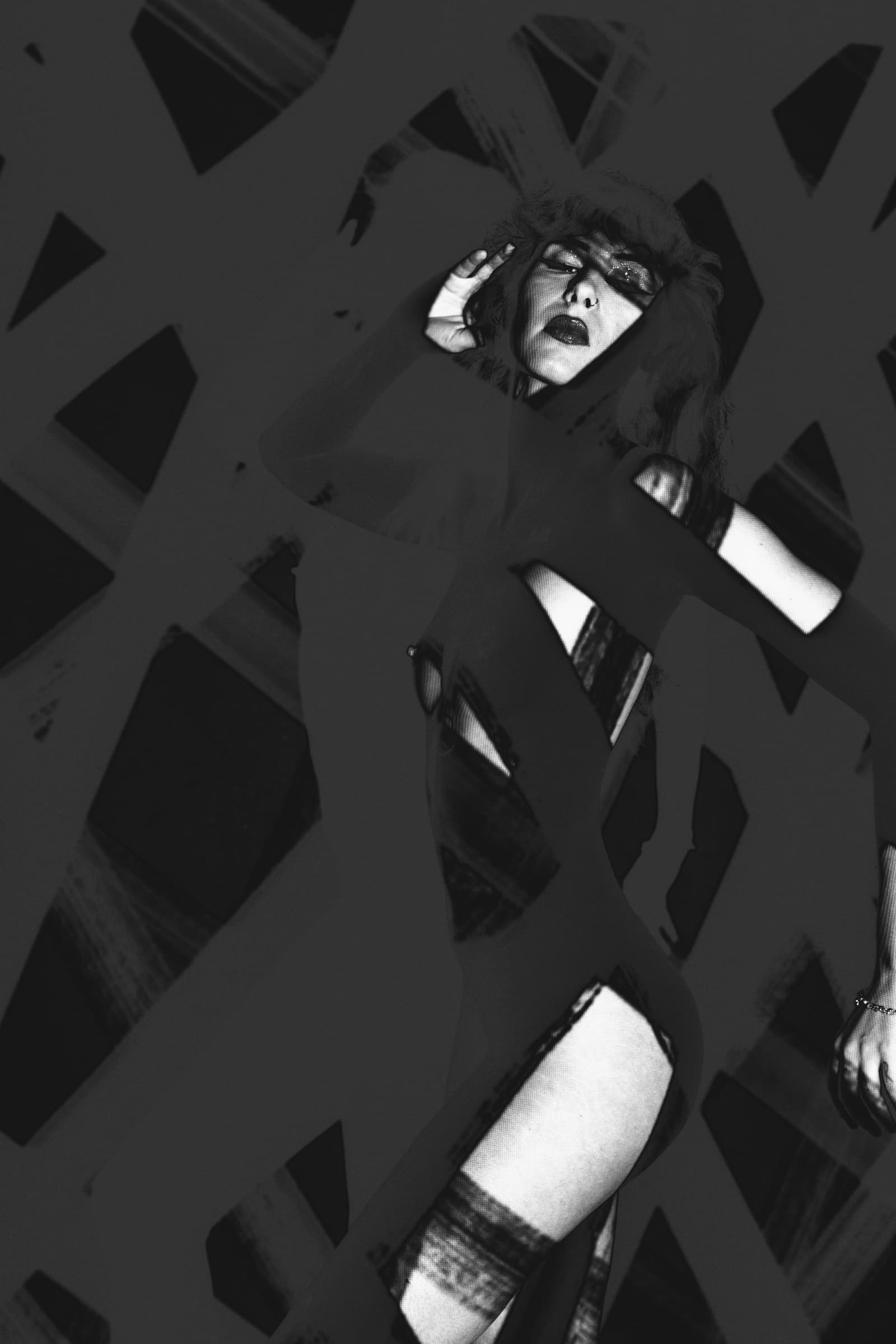 A black-and-white studio photograph of a person, partially obscured by a geometric, grid-like projection. The light is high-contrast, with dark solarised shadows cutting across the face and torso. The person is posed with one hand near their head, looking towards the camera with a serious expression.