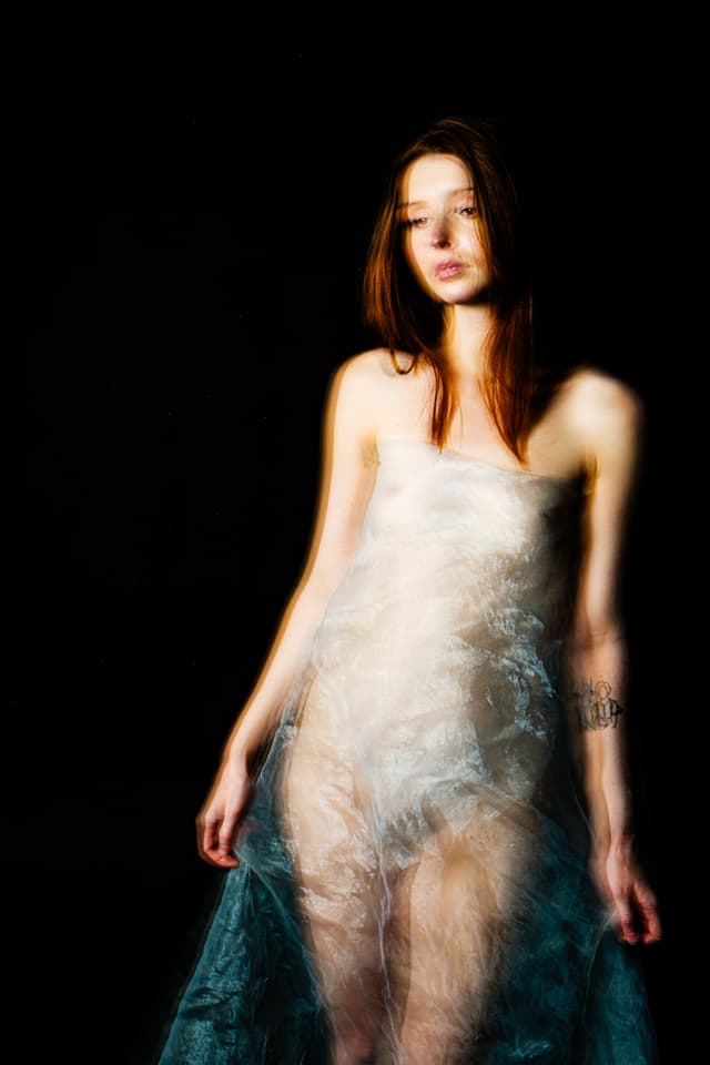 A person with auburn hair captured in a long-exposure shot against a pitch-black background, causing motion blur trails along their silhouette. They are wrapped in a single, sheer, translucent white fabric layer that partially obscures their form. Their head is tilted slightly to the side with eyes looking downward, and they have a dark tattoo visible on their inner forearm.