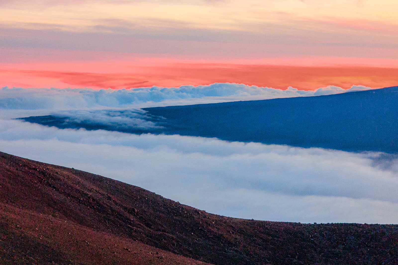 A panoramic view from high altitude looking over a thick blanket of clouds. In the foreground, a dark, reddish-brown volcanic slope angles sharply downward. In the middle ground, a long, dark ridge protrudes from the clouds. The sky above is filled with soft bands of orange and pink light during sunset.